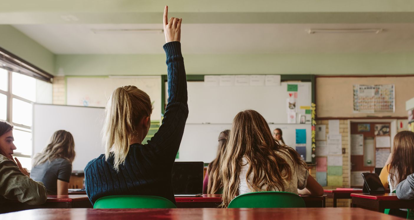 Rear view of female student sitting in the class and raising hand up to ask question during lecture. High school student raises hand and asks lecturer a question.