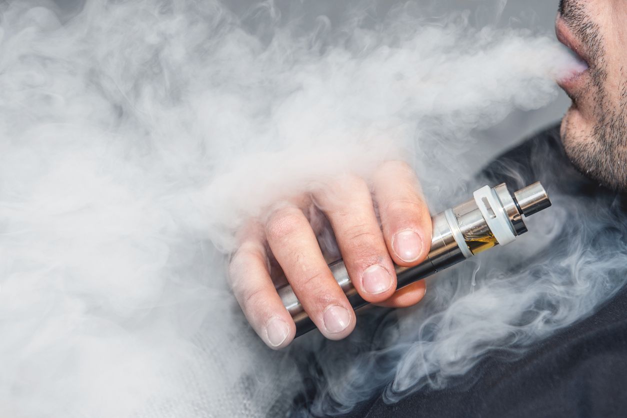 A man smokes an electronic cigarette on a gray background, blowing a stream of smoke. Copy space. Close up.