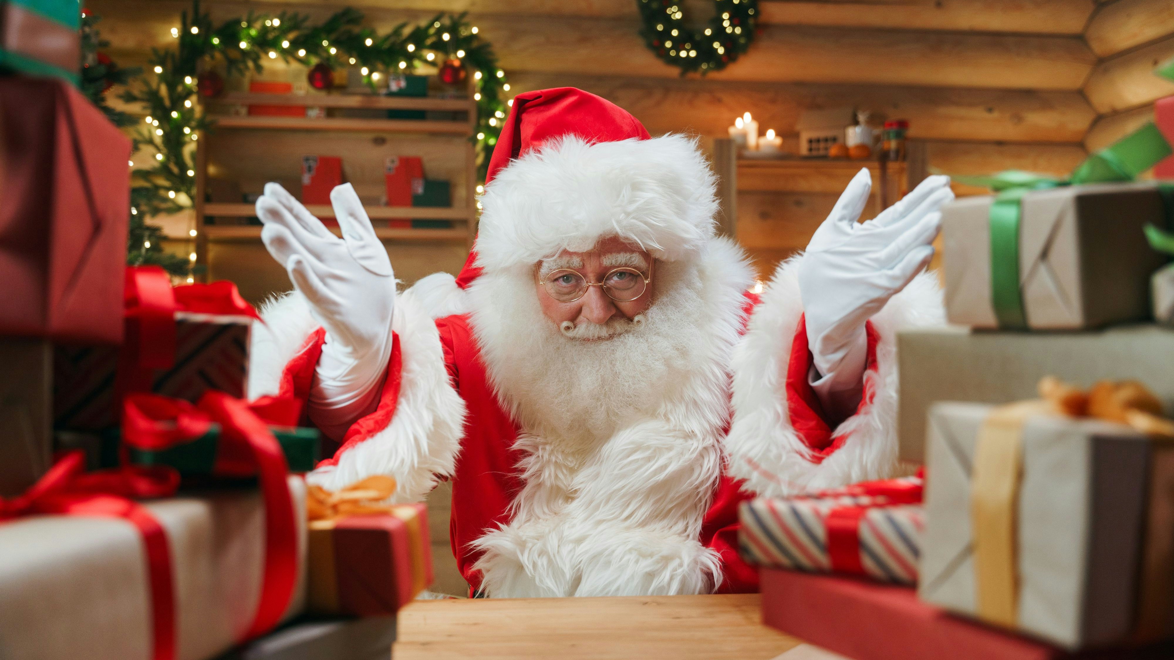 Santa Claus is  inviting us, by the gesture of his hands, we see  him in a centre of the photo  in the Christmas interior with a lot of presents in the foreground