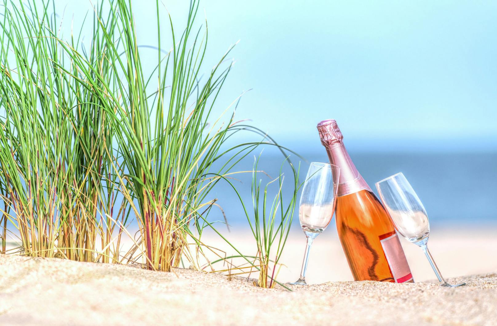 Bottle of rose colored champagne with drinking glasses on beach.