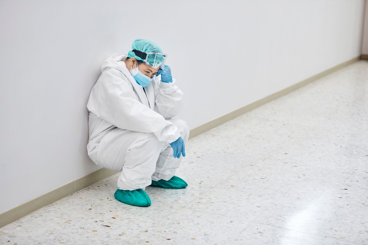 Tired healthcare worker crouching in corridor. Medical professional is exhausted in white coveralls. She is at hospital during COVID-19.