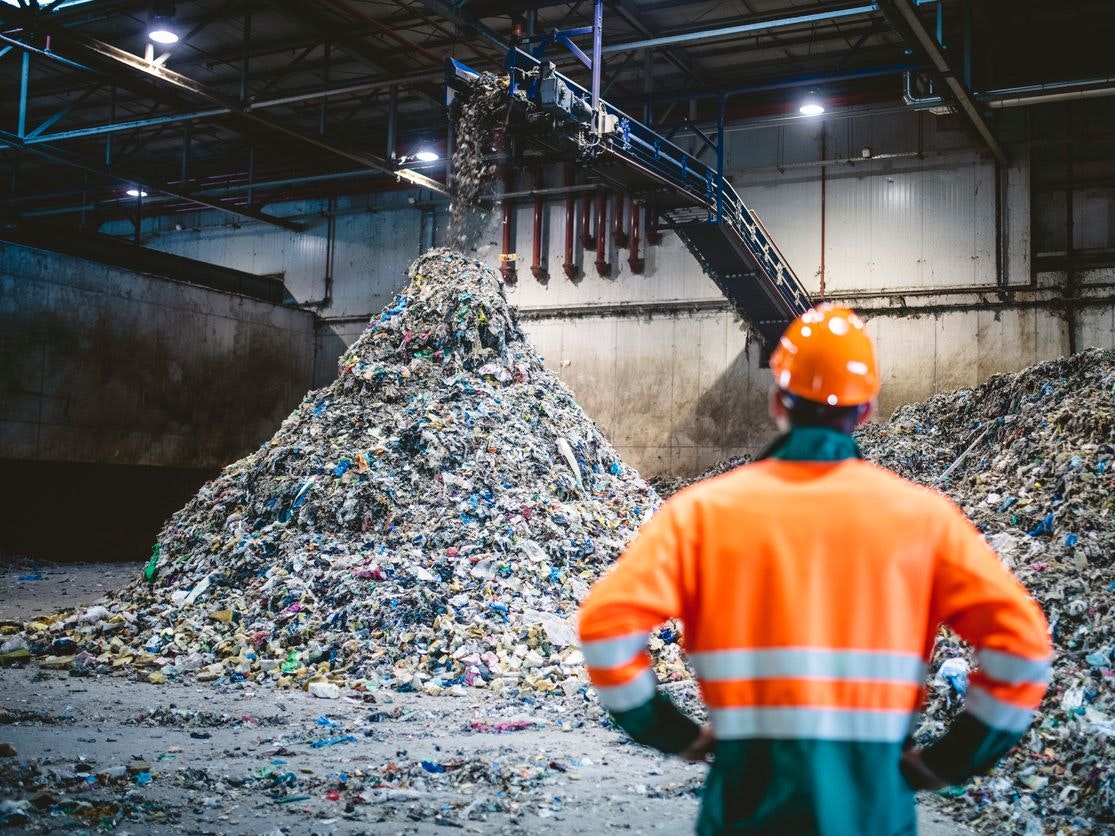 Rear view of young male worker in helmet, pollution mask, and reflective clothing observing waste falling from conveyor belt onto pile at facility.