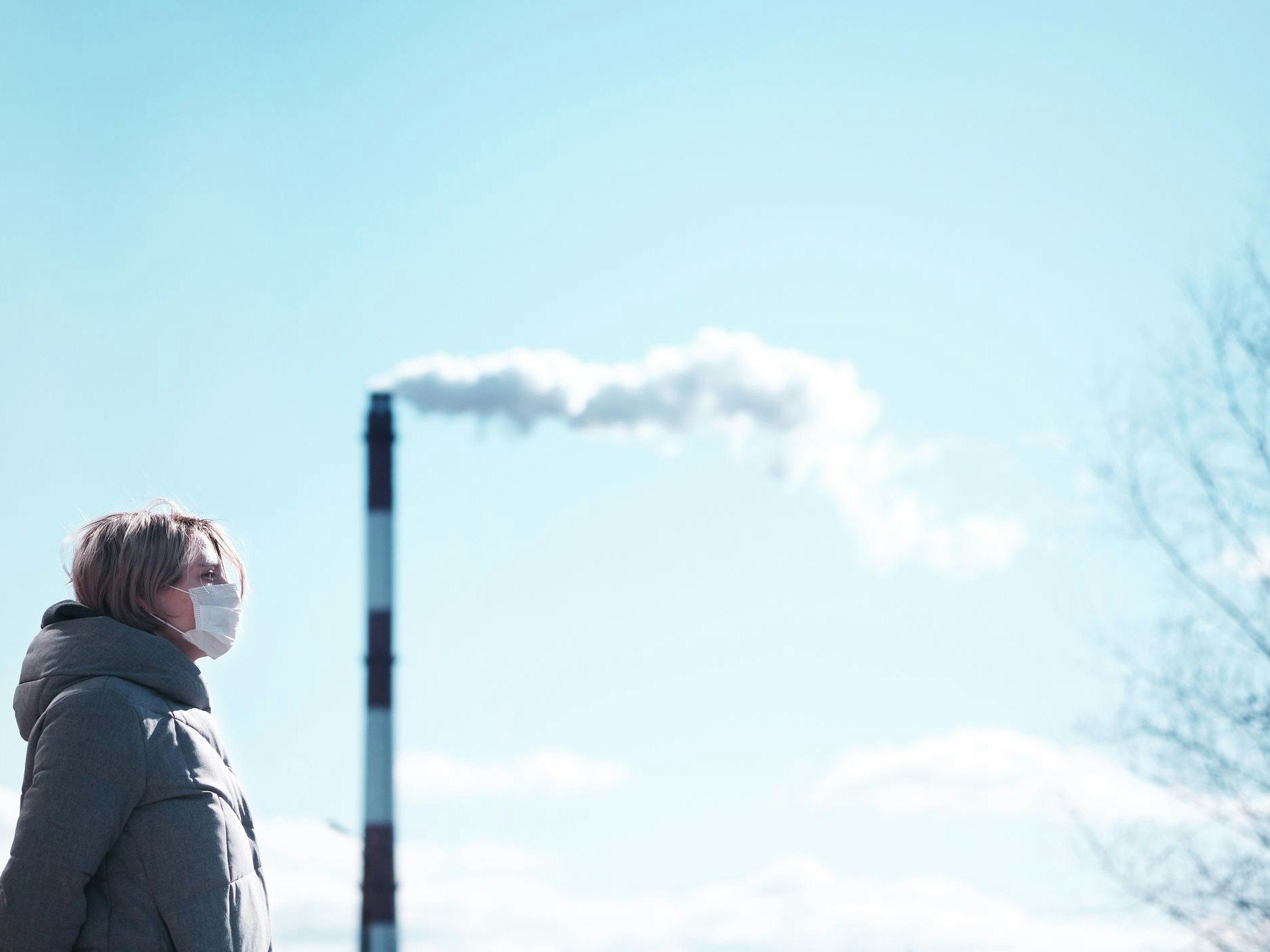 Young sad woman in a mask worried about environmental problems and air pollution is standing on the background of a pipe with smoke and thinking about the future