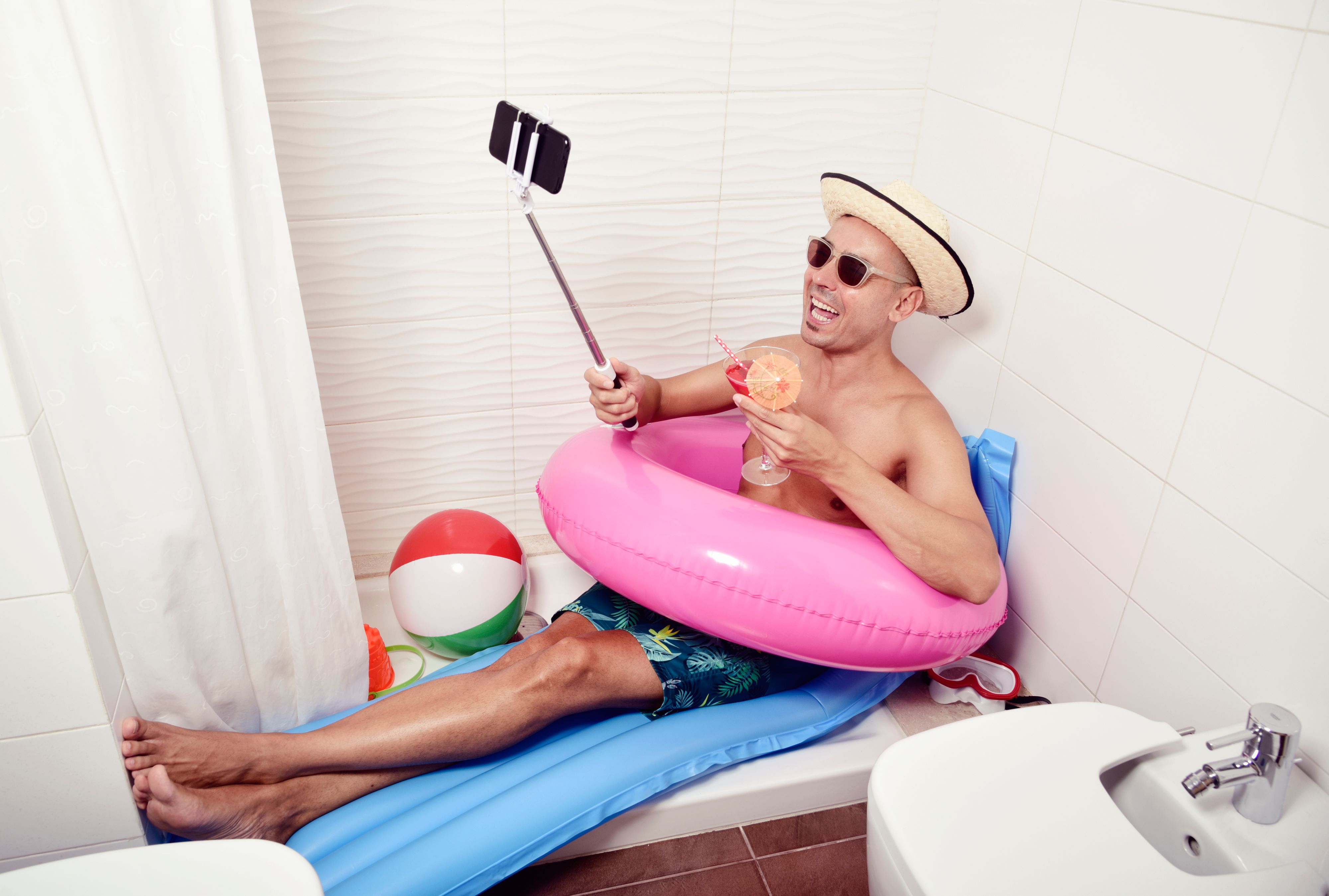a young caucasian man wearing sunglasses, a straw hat and a pink swim ring in a blue air mattress placed in the shower of a bathroom takes a selfie with his smartphone while is drinking a red cocktail