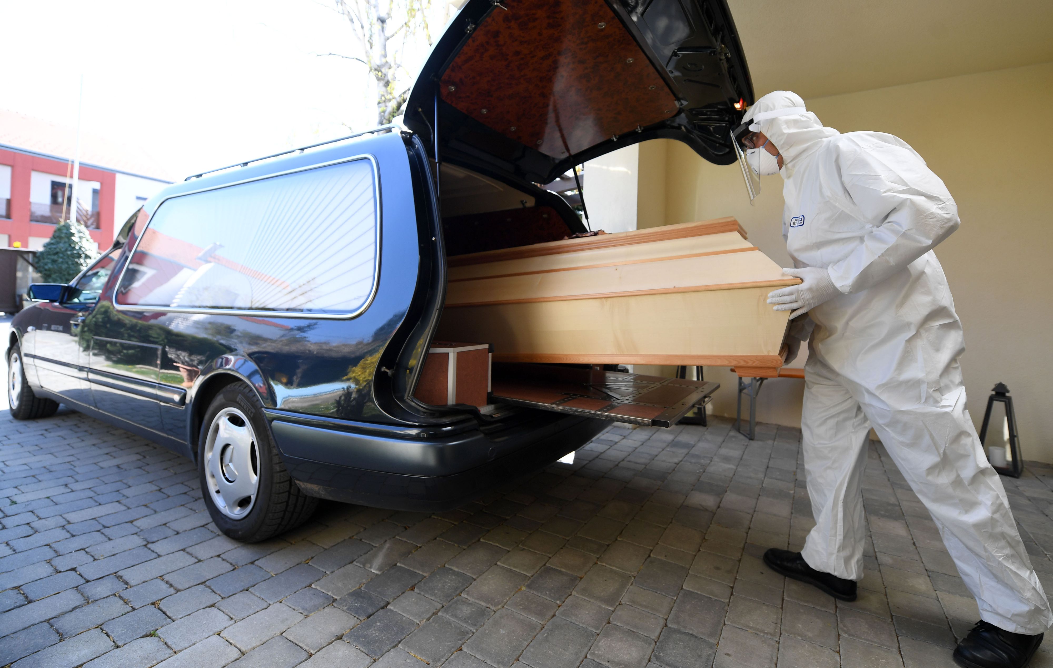 Ein Bestatter in Schutzkleidung schiebt einen Sarg in einen Leichenwagen. Symbolbild