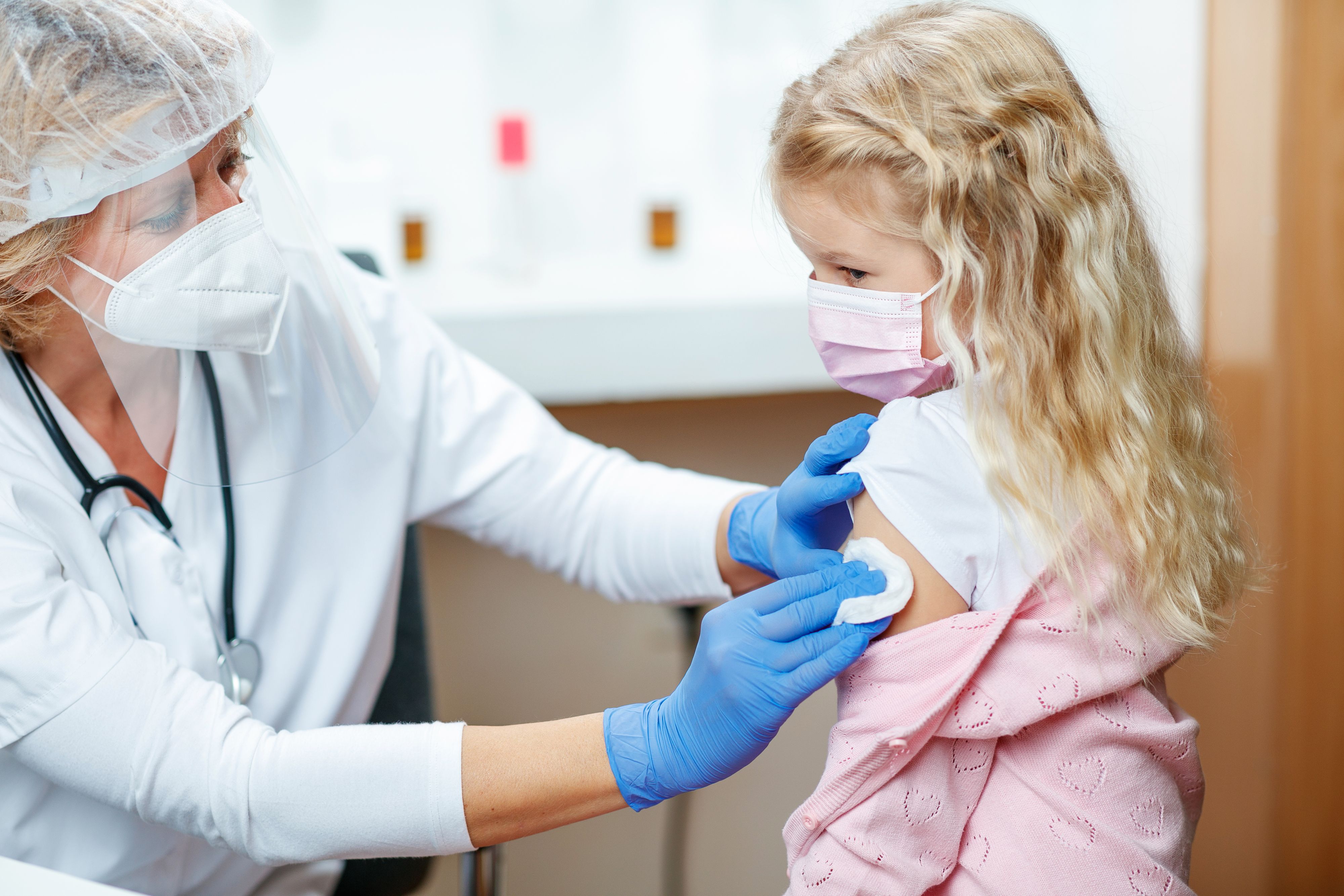 Female doctor in full protective workwear cleaning girl's arm before injecting COVID-19 vaccine, girl wearing protective face mask