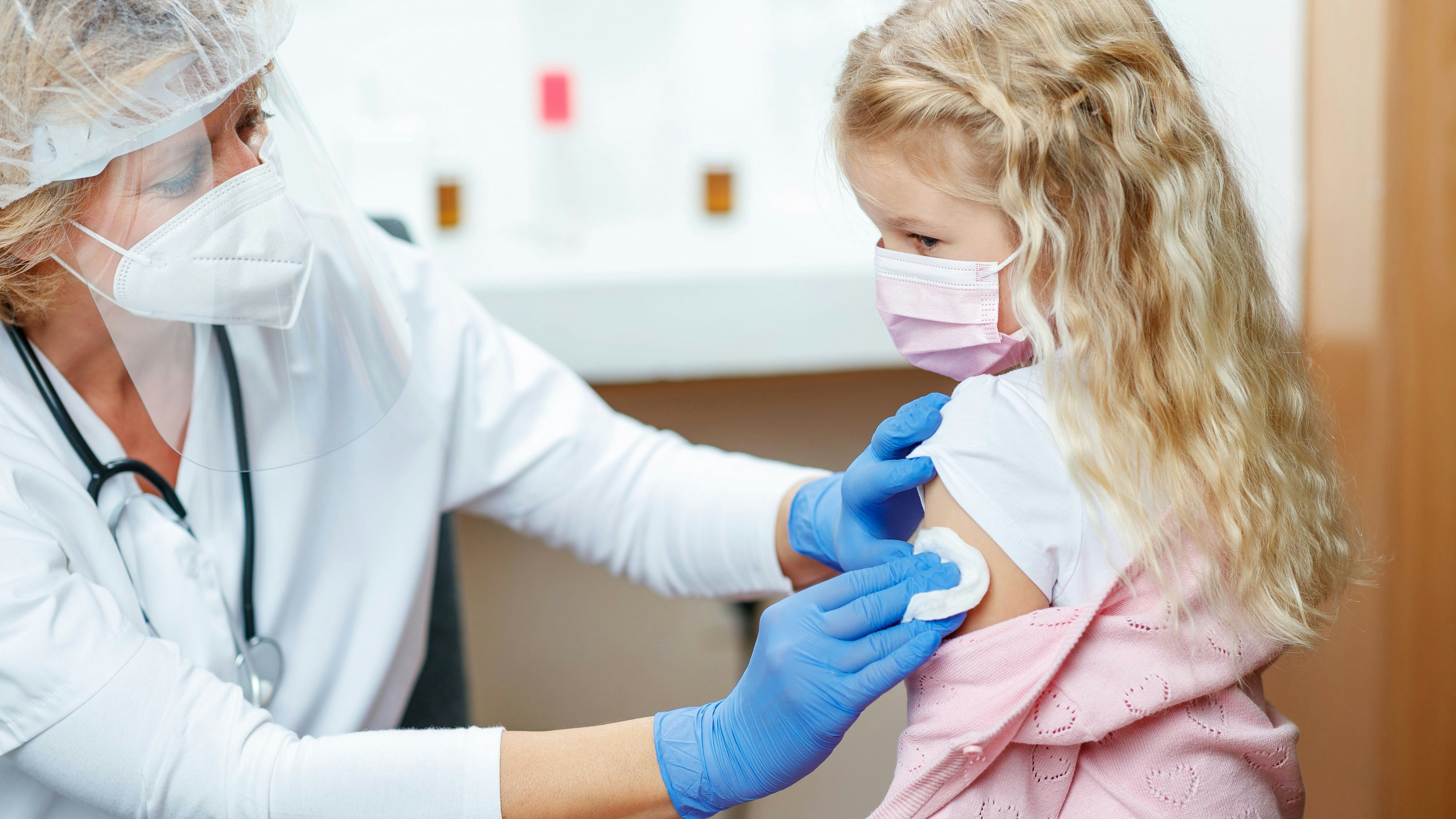 Female doctor in full protective workwear cleaning girl's arm before injecting COVID-19 vaccine, girl wearing protective face mask
