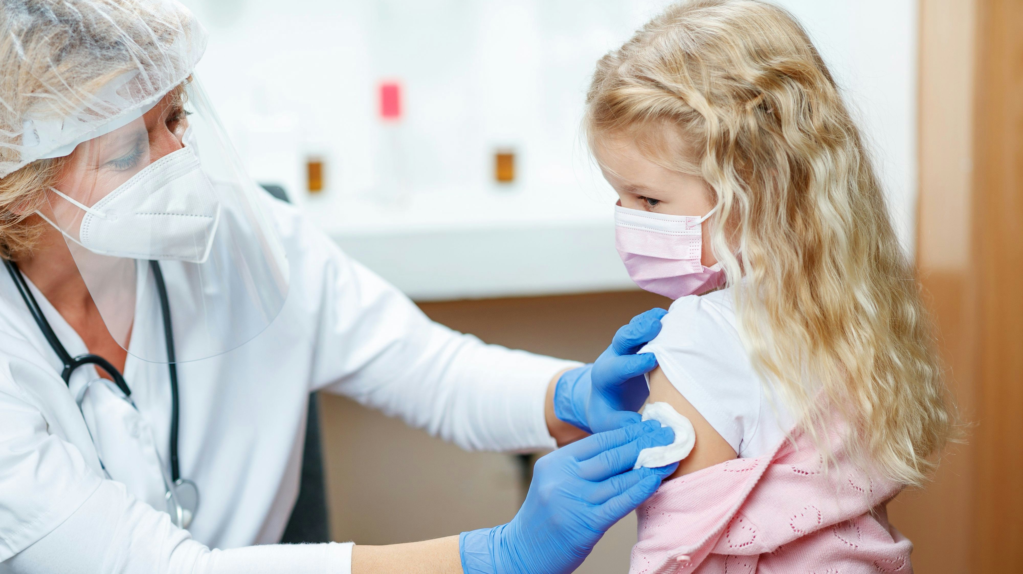 Female doctor in full protective workwear cleaning girl's arm before injecting COVID-19 vaccine, girl wearing protective face mask