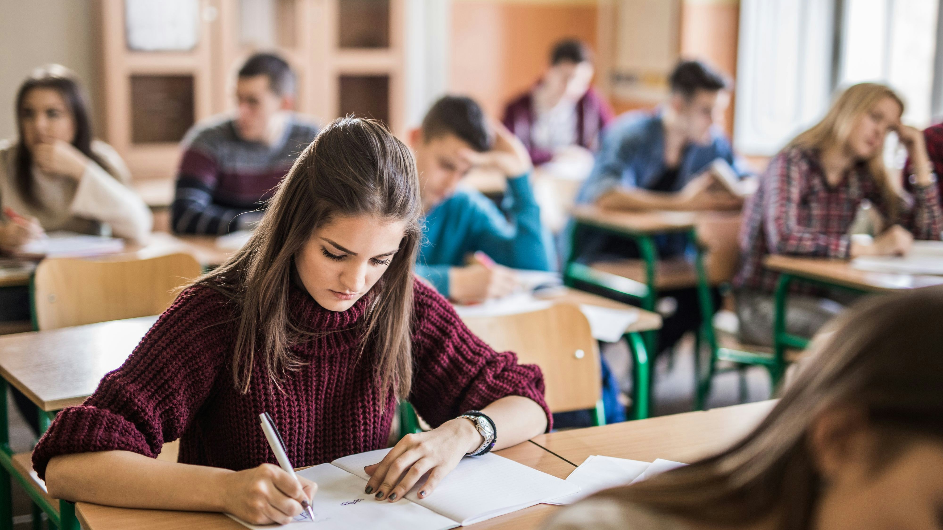 Teenage girl taking notes while sitting in the classroom with her classmates.