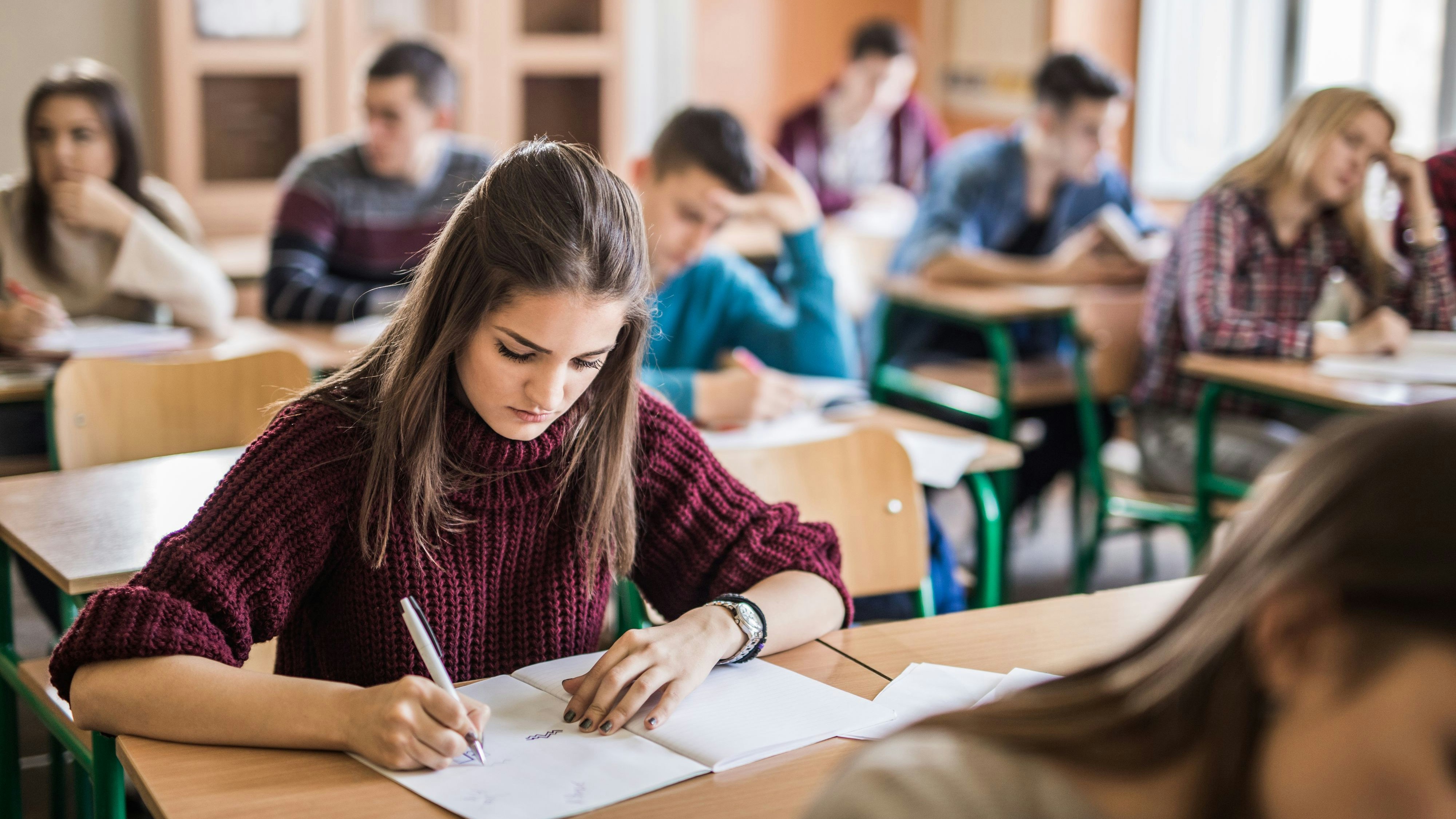 Teenage girl taking notes while sitting in the classroom with her classmates.