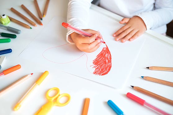 Close up of child's hands drawing red heart at white paper within colorful pens and pencils.