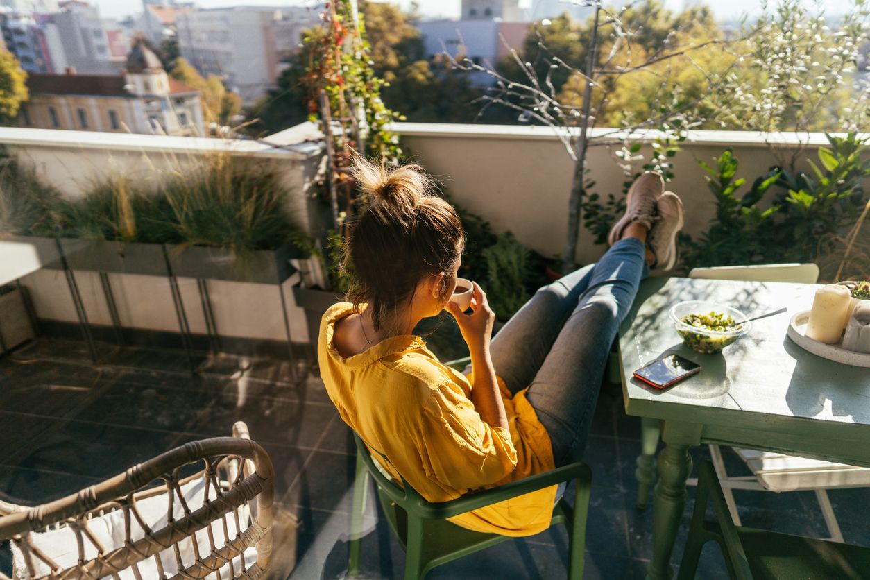 Photo of a woman drinking first morning coffee, eating 'take out' food and reading daily news online - on the balcony of her apartment while enjoying springtime sun