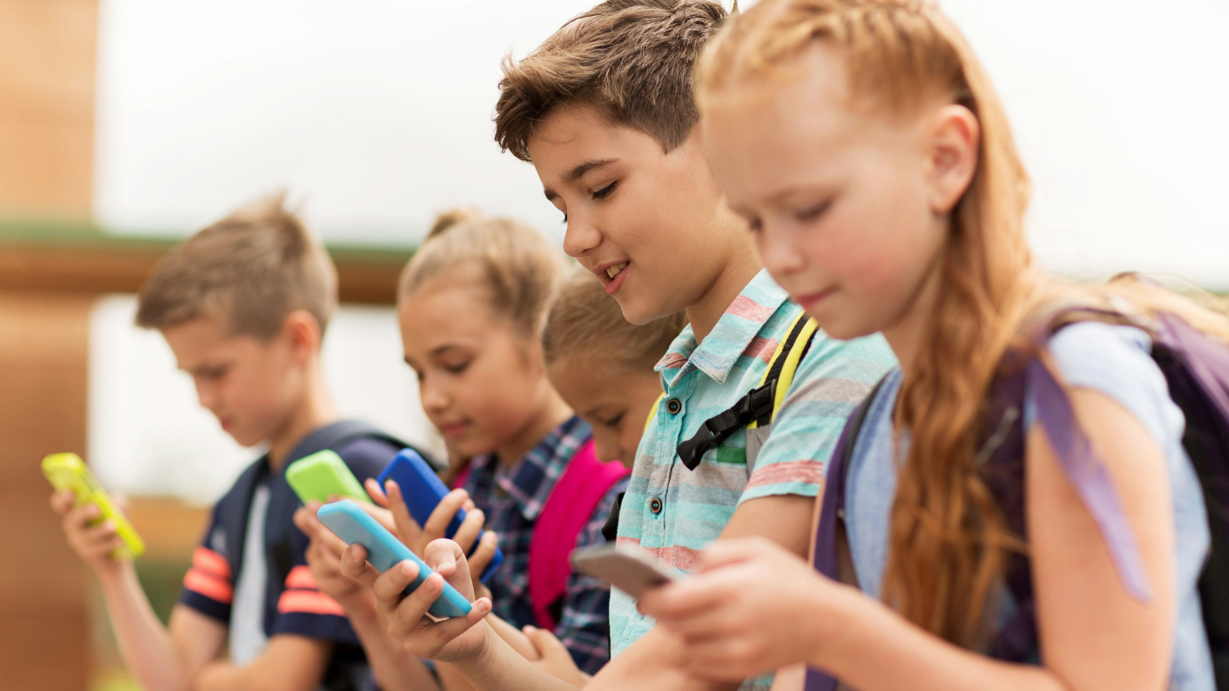 primary education, friendship, childhood, technology and people concept - group of happy elementary school students with smartphones and backpacks sitting on bench outdoors