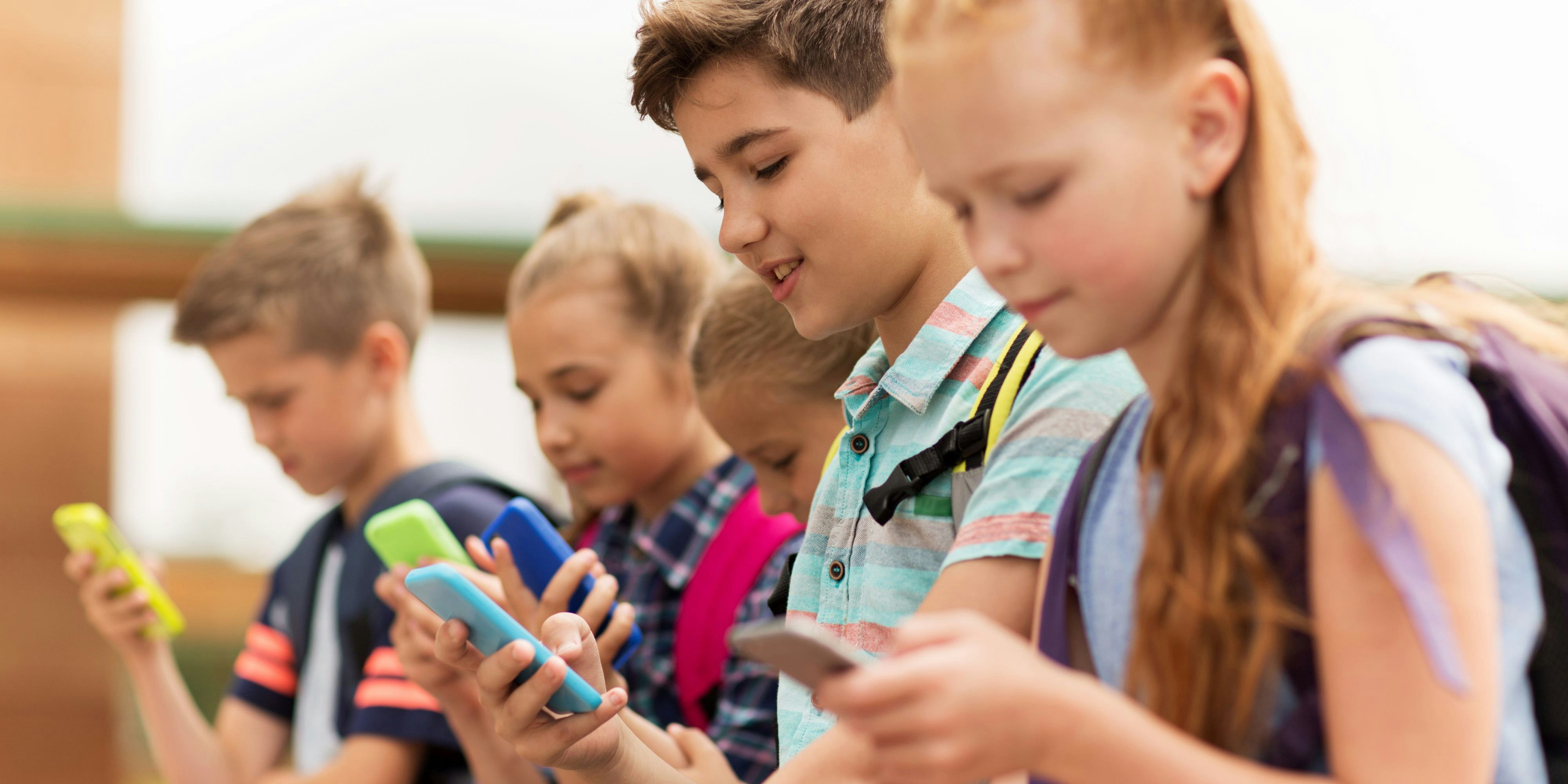 primary education, friendship, childhood, technology and people concept - group of happy elementary school students with smartphones and backpacks sitting on bench outdoors