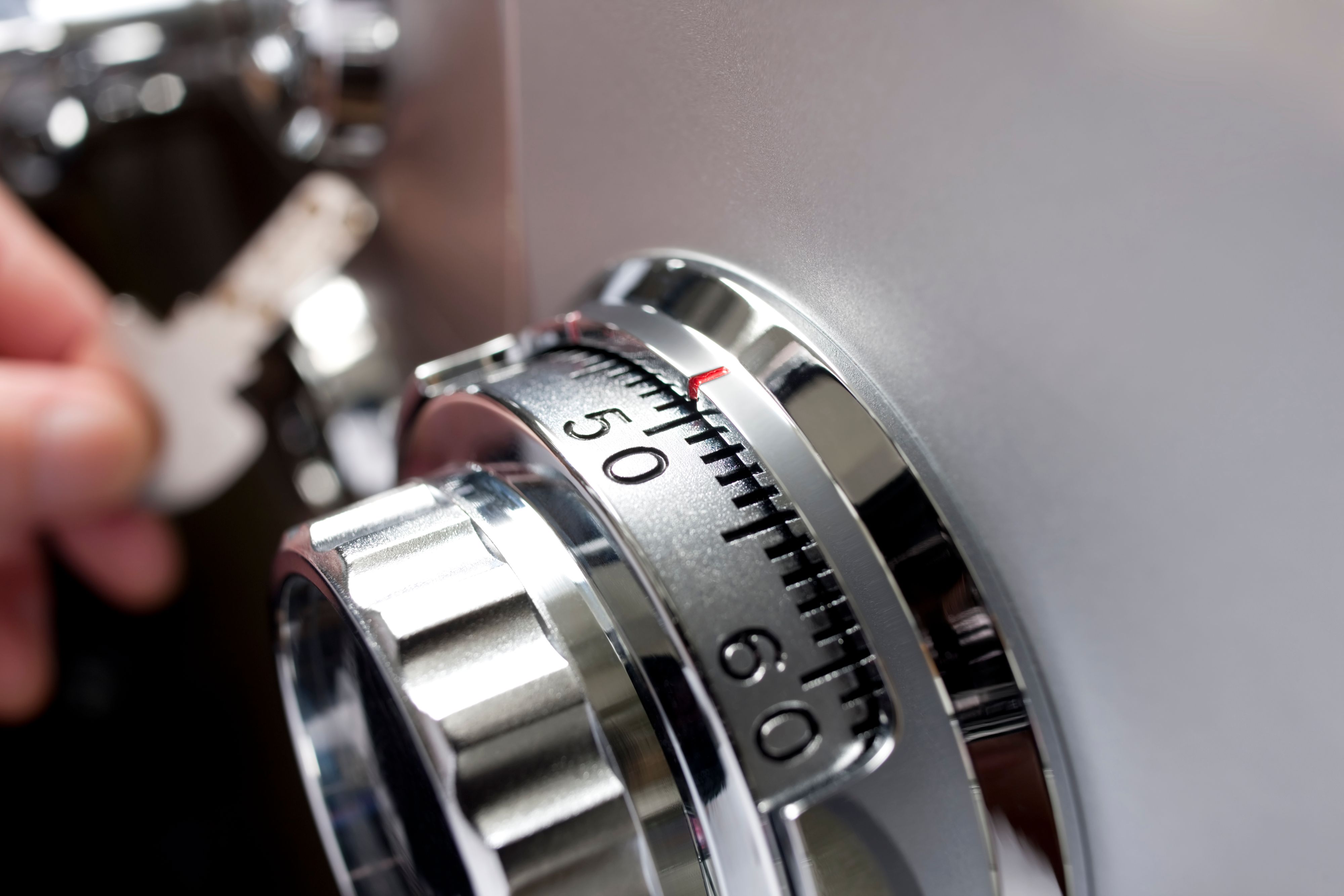 The dial of an office or home safe. Shallow depth of field. Focus on the centre of dial, an out of focus hand with one of the locking safe keys in the background. 