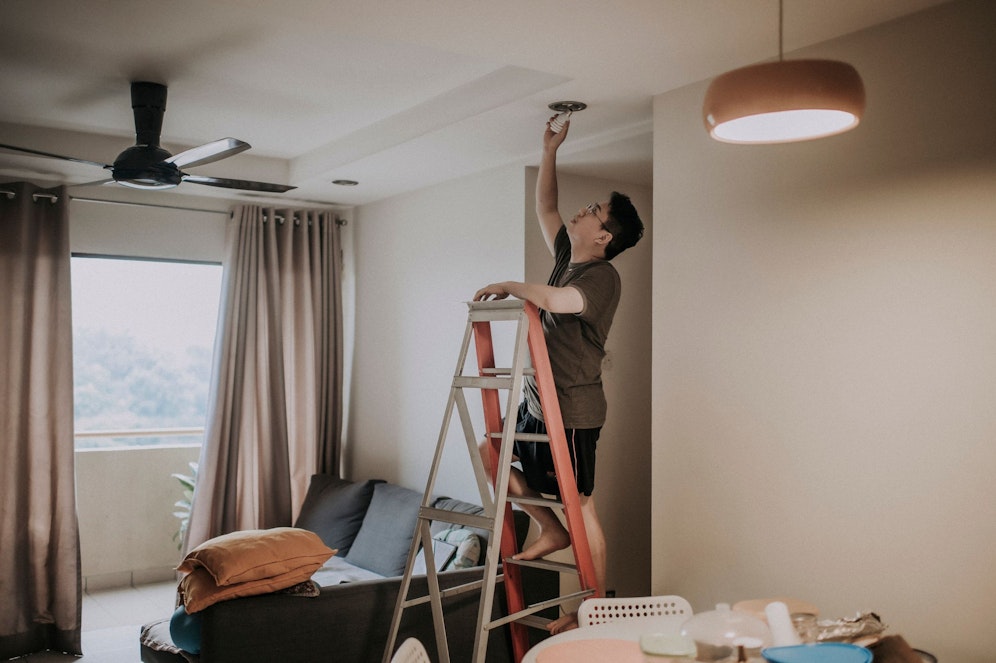 Ein Mann steht auf einer Leiter, um eine Glühbirne zu wechseln. Symbolbild