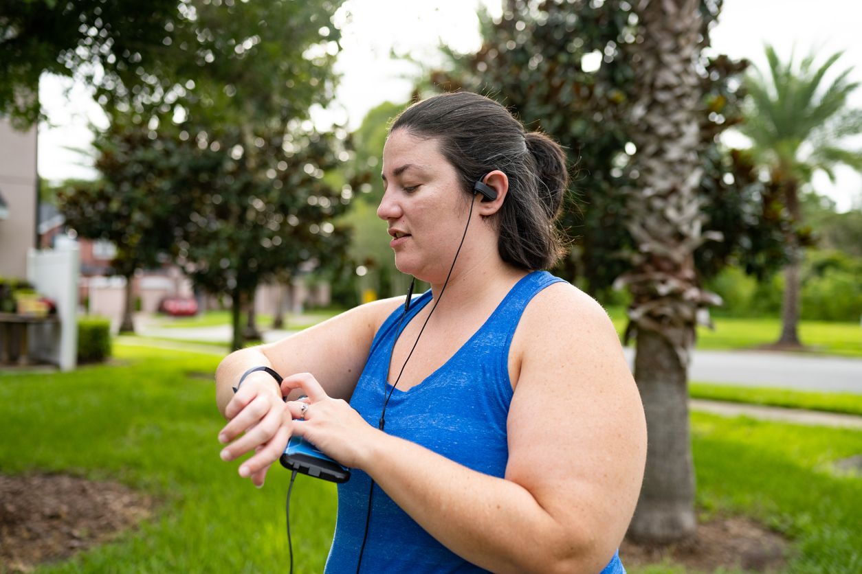 This is a photograph of a body positive Caucasian woman in her 30’s checking her fitness tracker while outdoors on a walk in a suburban Orlando, Florida neighborhood.