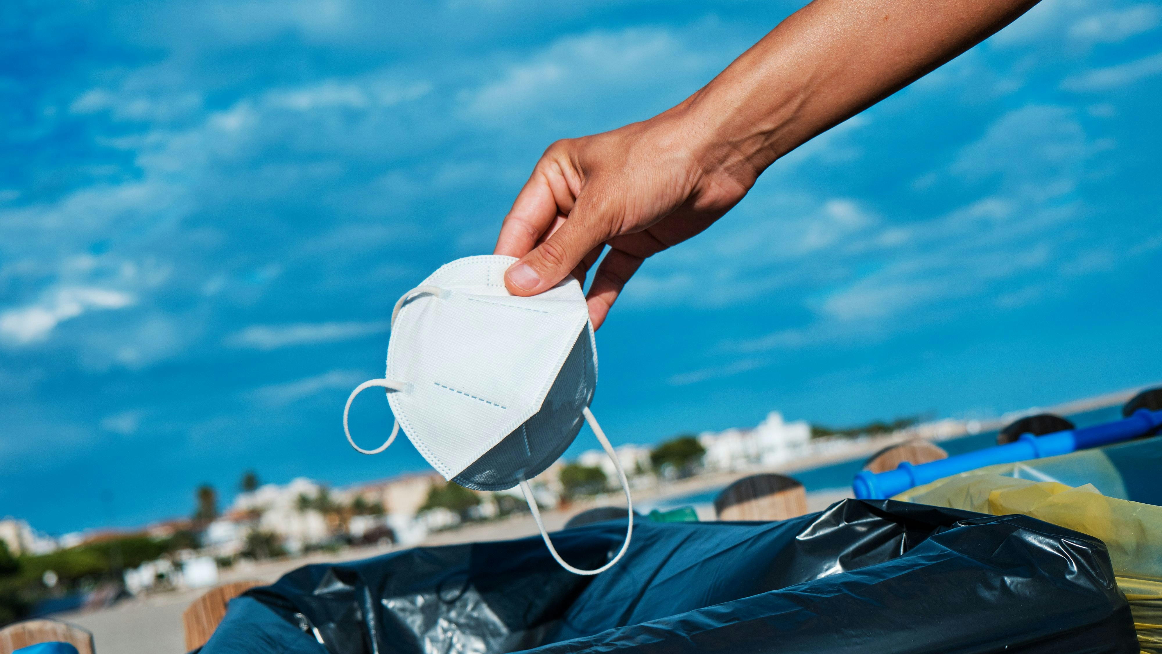 closeup of a caucasian man throwing a used white disposable face mask to the trash next to the beach