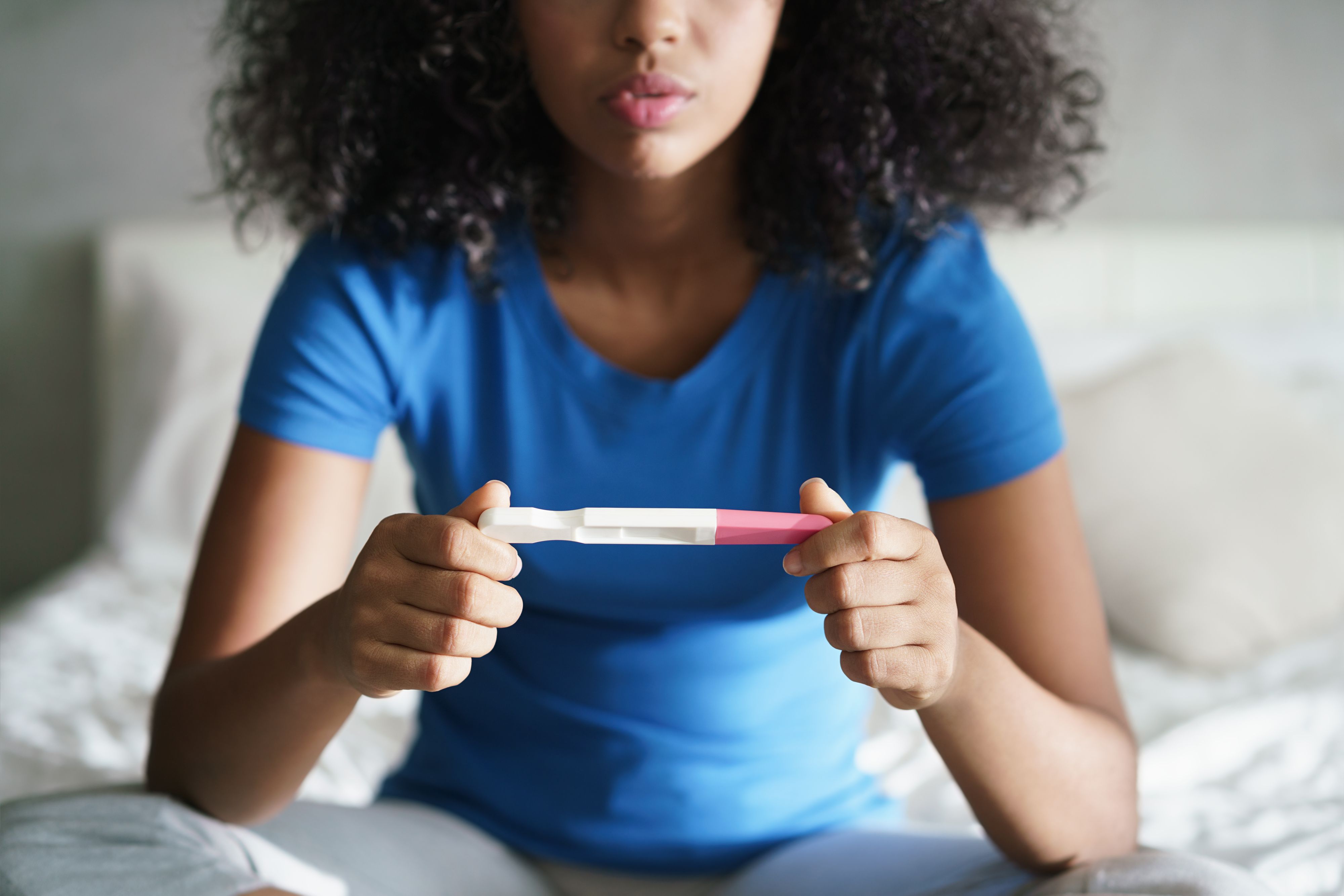 Disappointed hispanic girl getting unexpected result from pregnancy test kit. Sad young latina woman sitting alone on her bed.