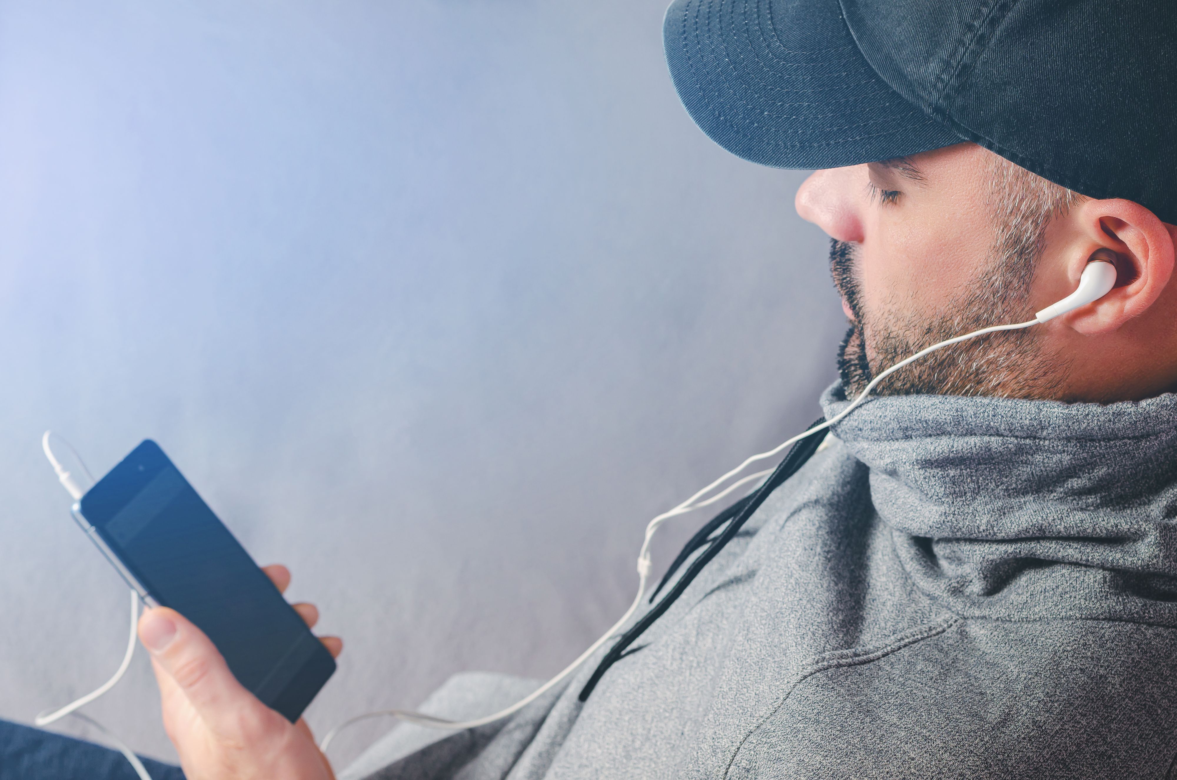 Cropped portrait of bearded man in black cap watching videos or listening to the music with earphones on smartphone. Copy space