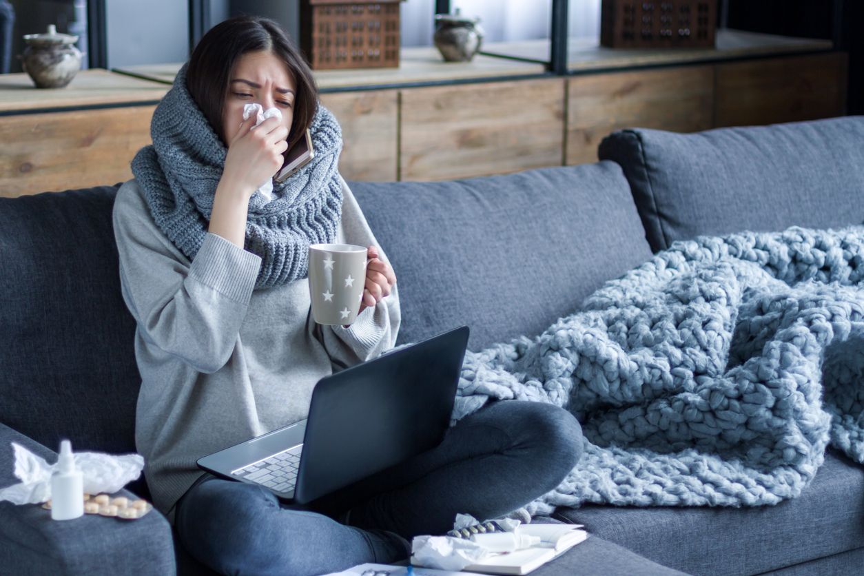 Ill brunette girl is sitting at sofa and working remotely on laptop. Female is blowing out snot, having fever and headache. Young woman is treated at home, took sick leave. Winter cold and flu concept