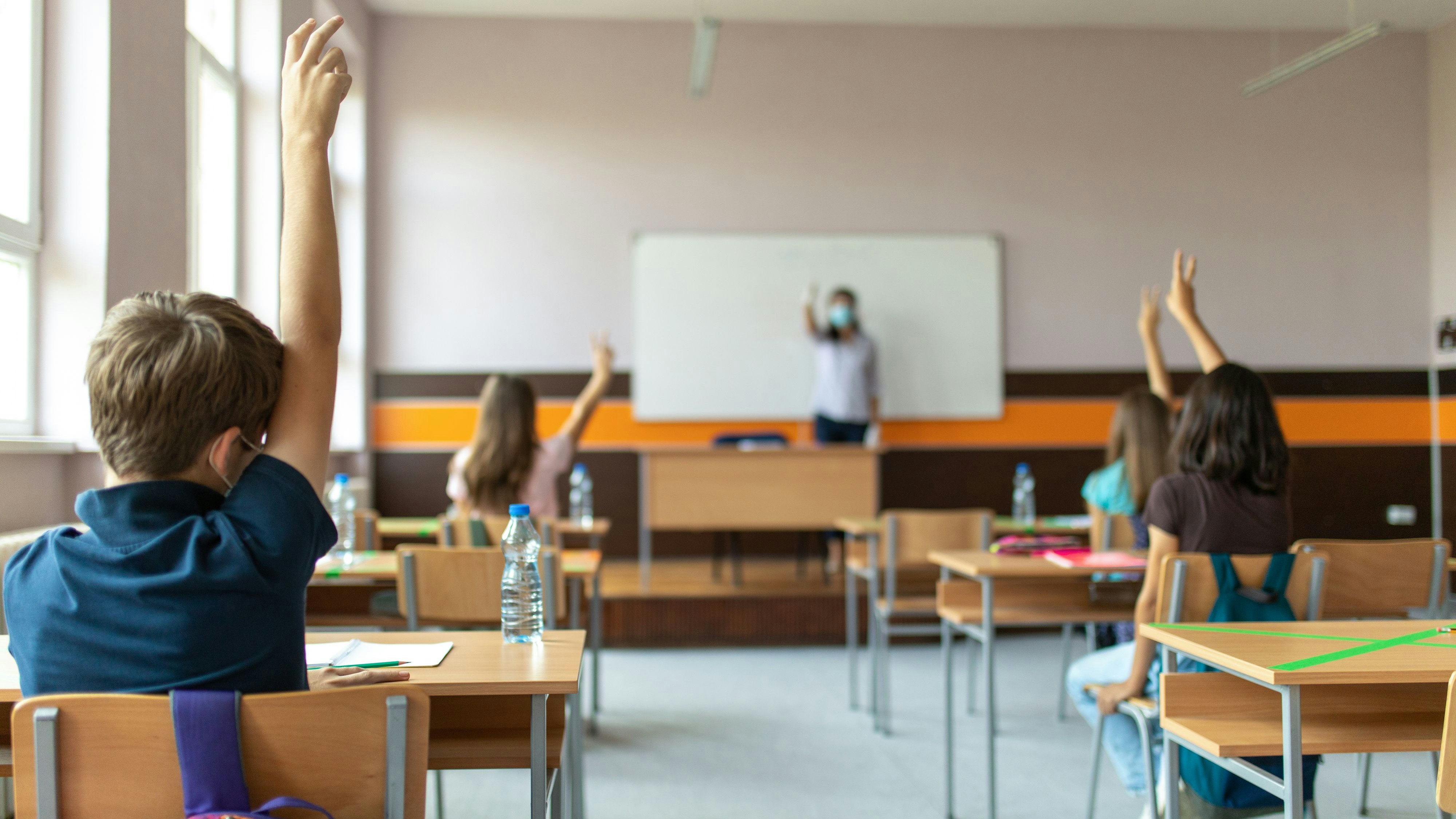 Students with protective masks sitting in school desks in their classroom. School desks are marked with a cross to mark a place where sitting is not allowed to maintain social distance during corona virus .