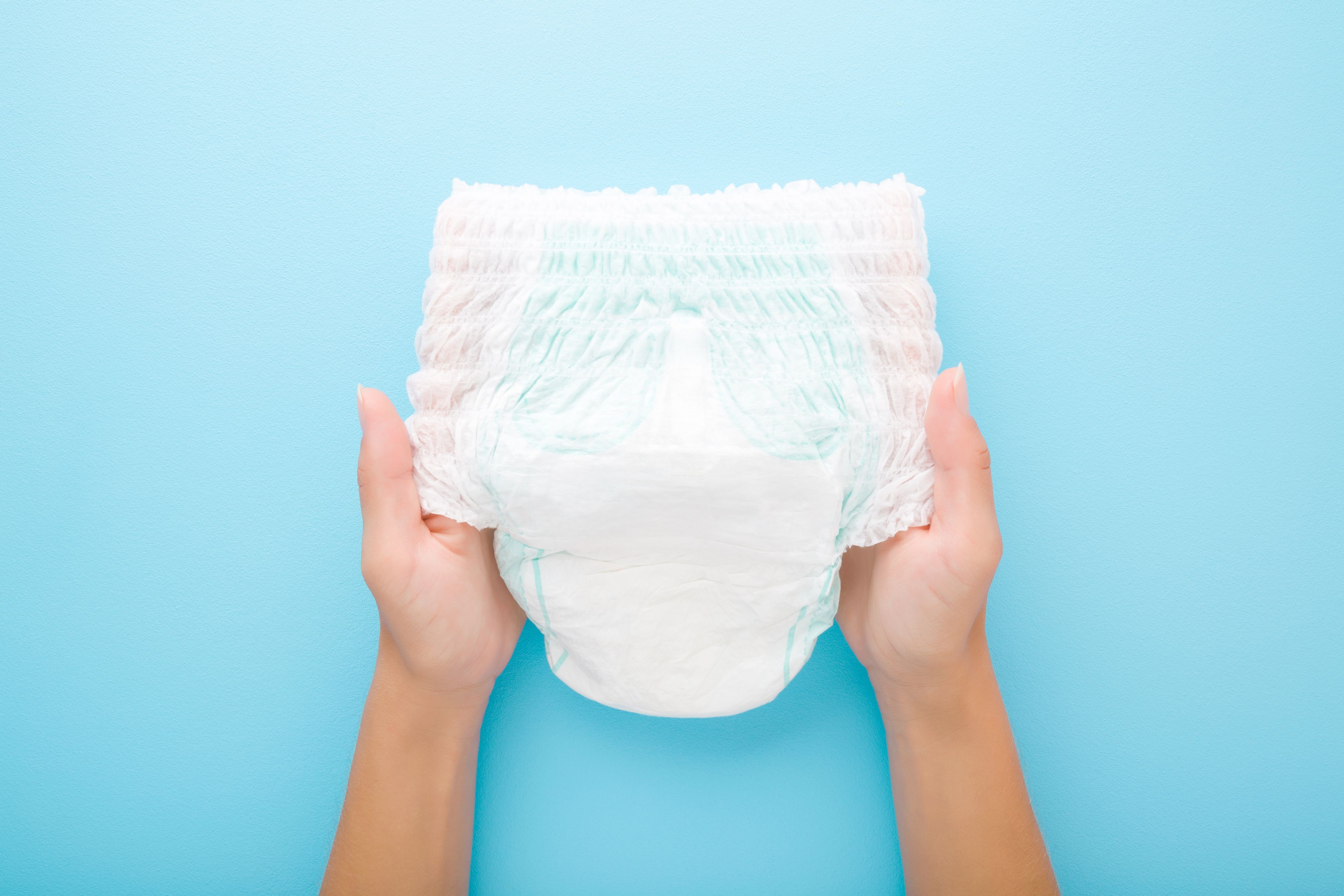 Young mother hands holding white baby diaper pants on light blue table background. Pastel color. Closeup. Point of view shot. Top down view.
