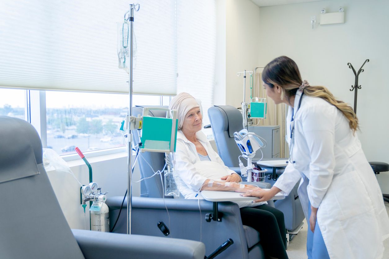 A senior woman is sitting on a chair in a chemotherapy treatment room. She is wearing a headscarf due to hair loss. Her female oncologist is checking in on her and providing her with moral encouragement.