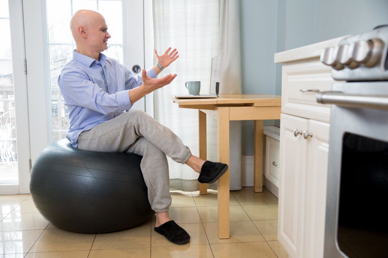 Man wears a dress shirt and jogging pants and sits on a yoga ball while on a business work from home zoom call on the laptop