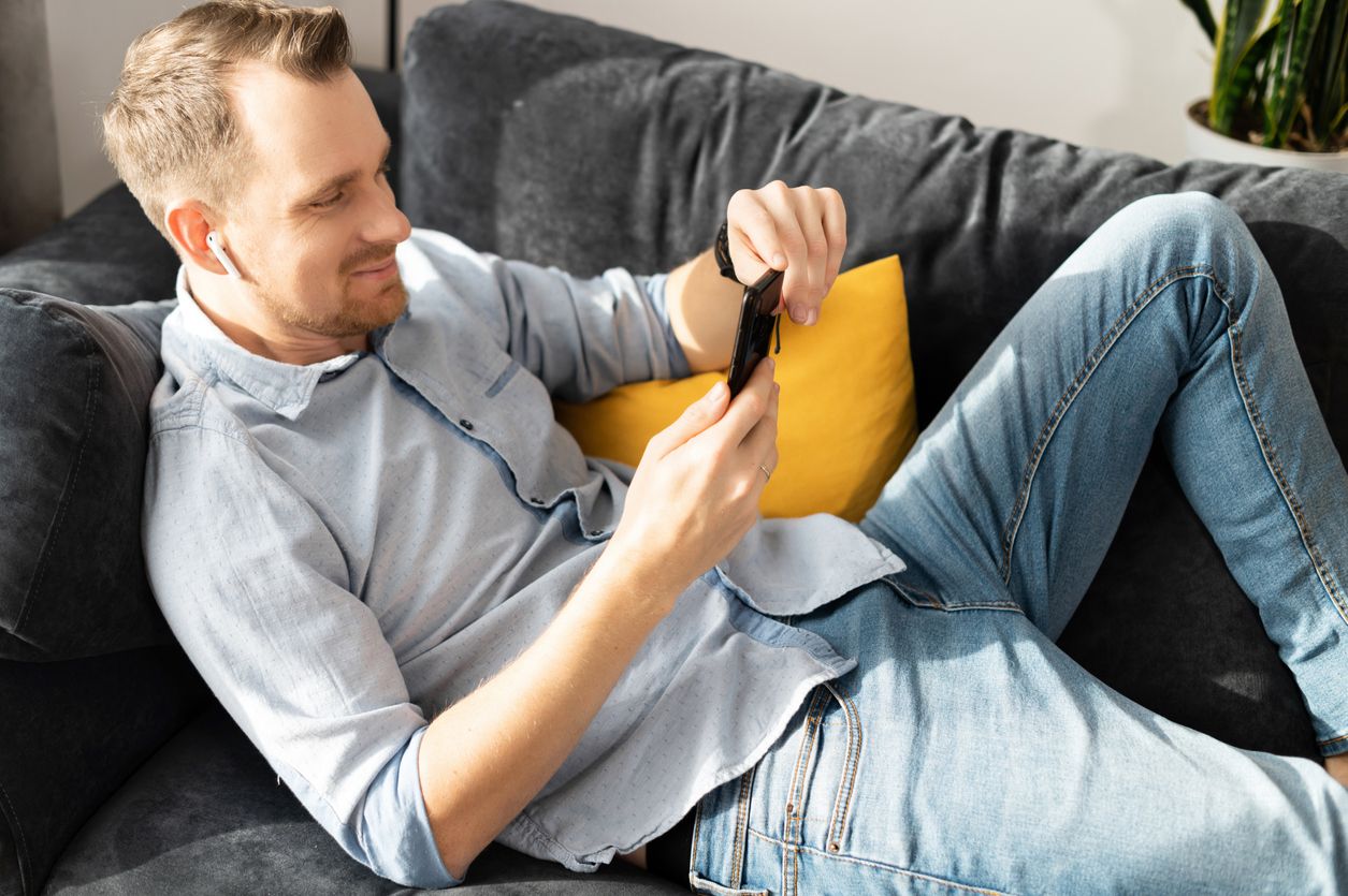 Smiling cheerful guy is lying on the couch and relax with a smartphone in hands. A young man in smart casual wear spends time with a gadget at home