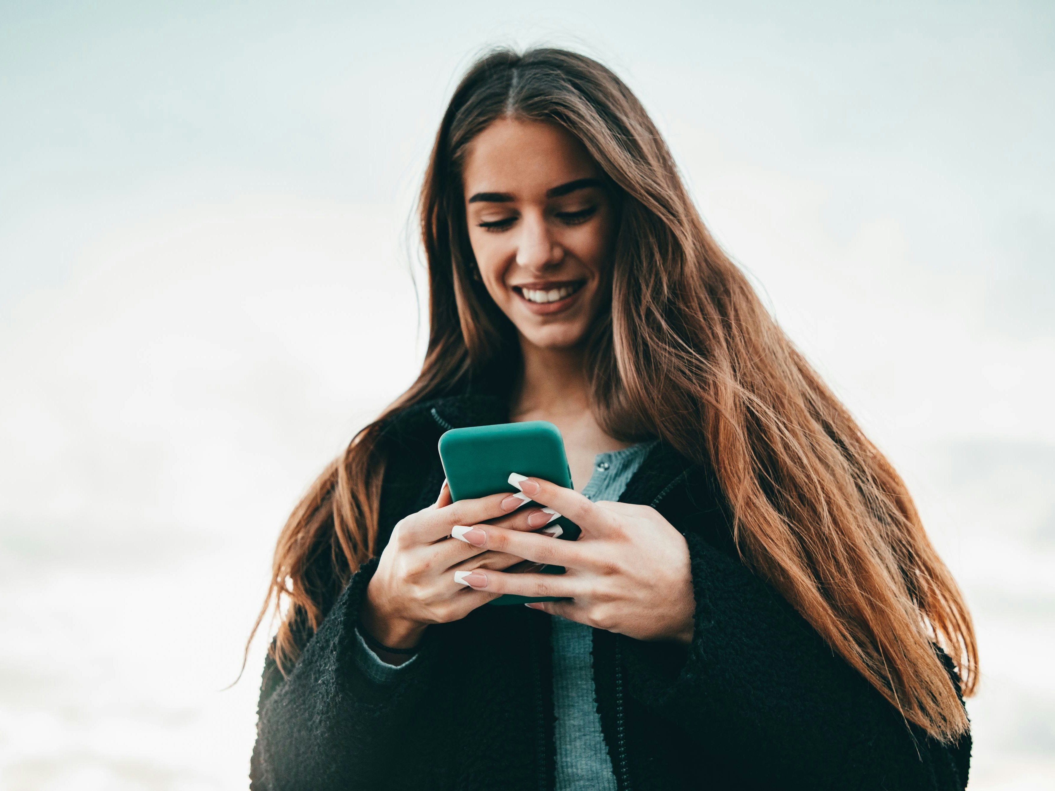 Young cheerful smiling happy woman checking messages on her smart phone outdoors in wintertime, Real People Outdoor Millenial Youth Lifestyle Portrait.