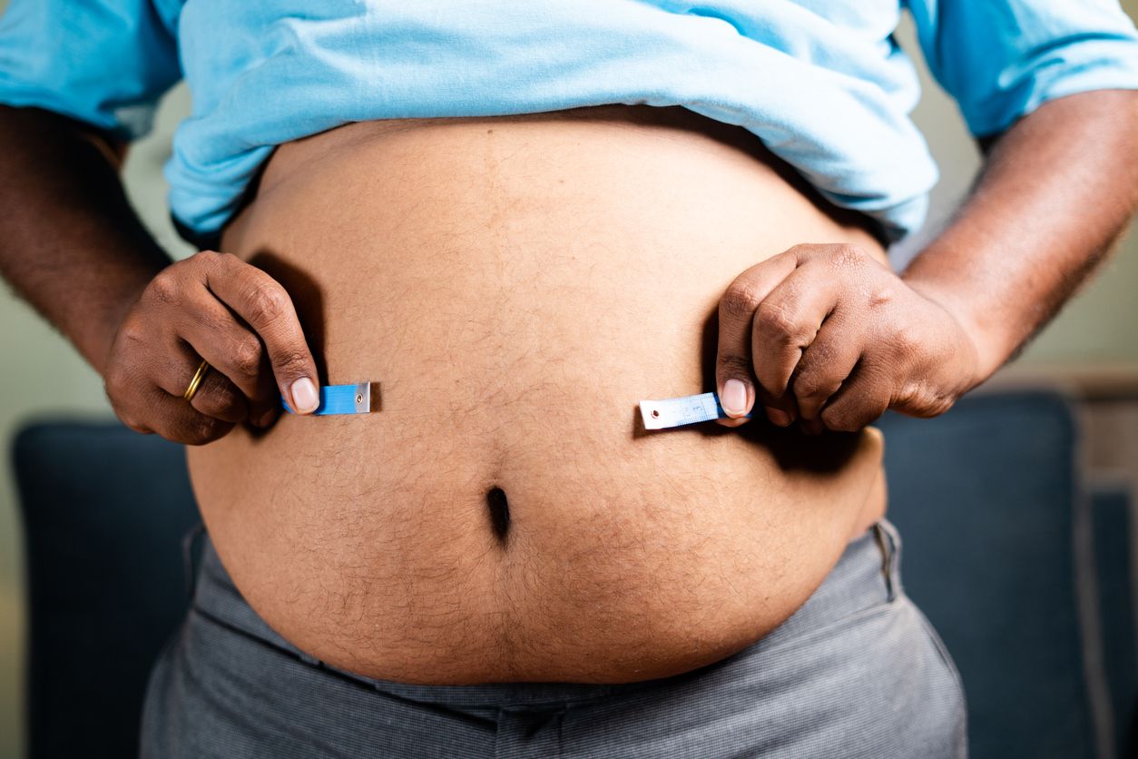 Unrecognizable unhealthy obese man checking stomach or waist fat using measuring tape before diet at home - concept of suffering body overweight due to junk food habits