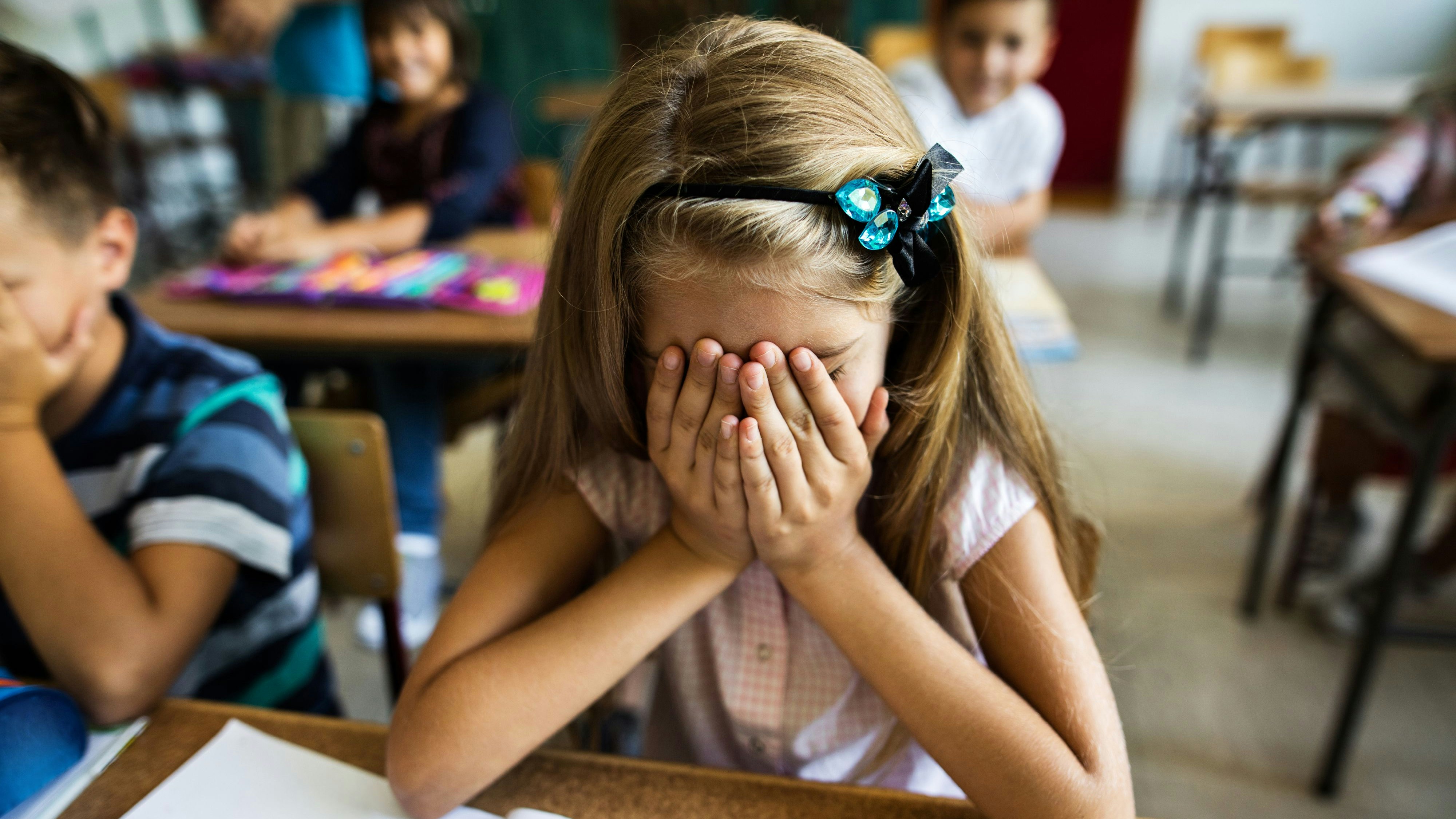 Little girl in the classroom feeling stressed and covering her face with hands while sitting at the desk.