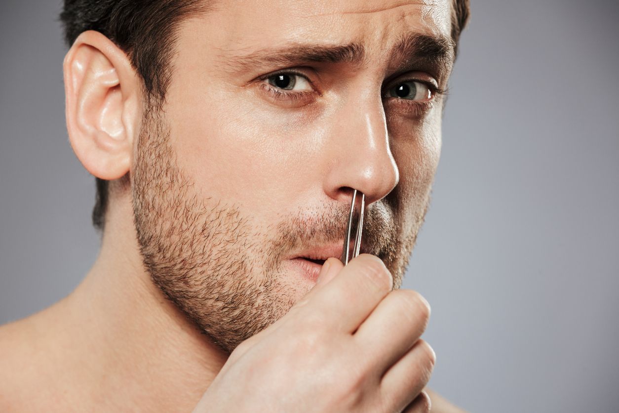Close up of terrifoed man removing nose hair with tweezers isolated over gray background