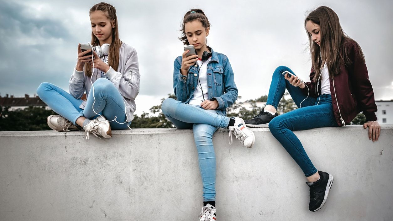 three teenage girls sitting outdoors on concrete wall and looking at their smartphones