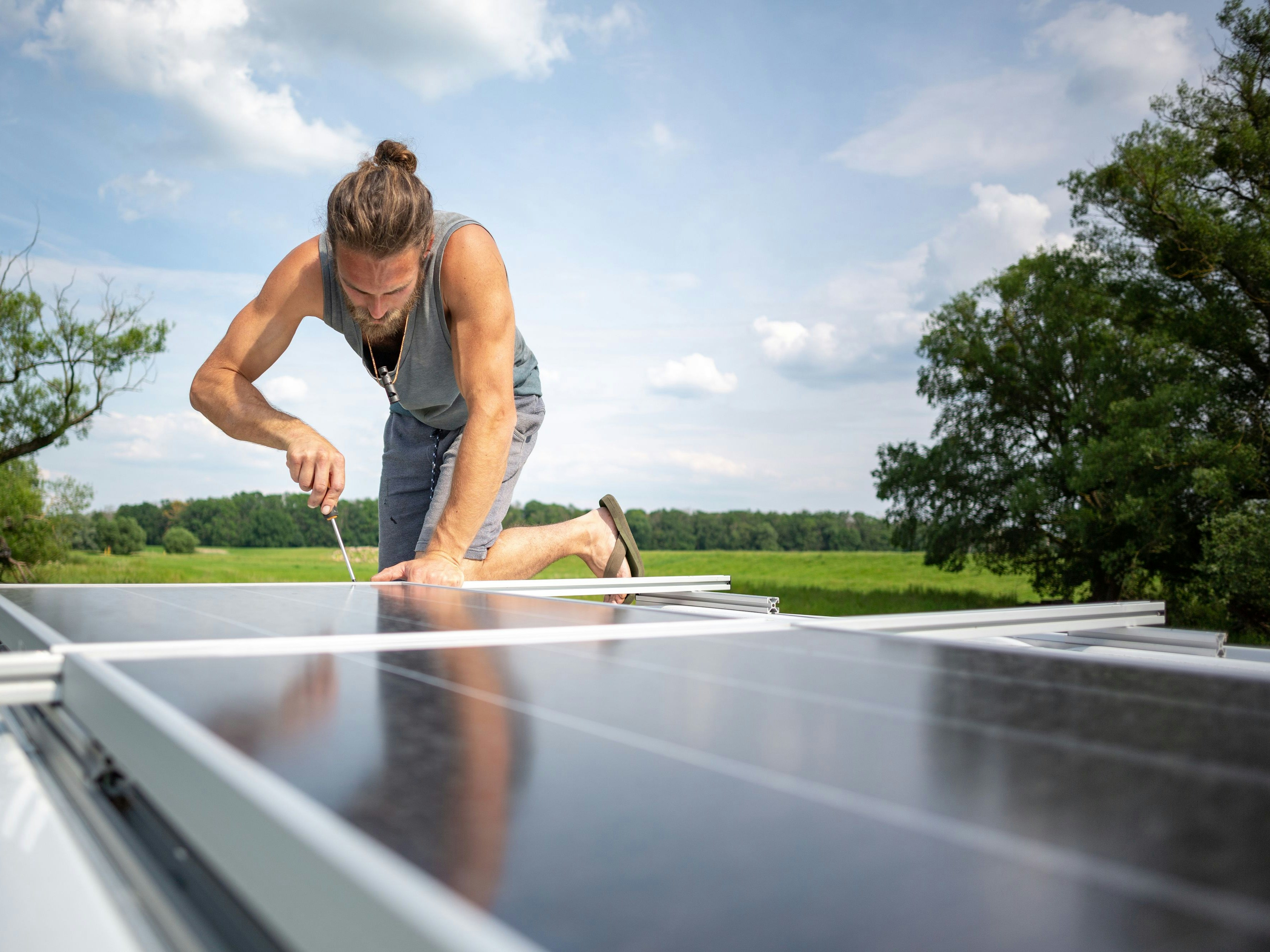 Young man on top of the roof of a camper van with solar panels. He is using a screwdriver to work on the panels. The sun is shining and a green meadow with trees is visible in the background.