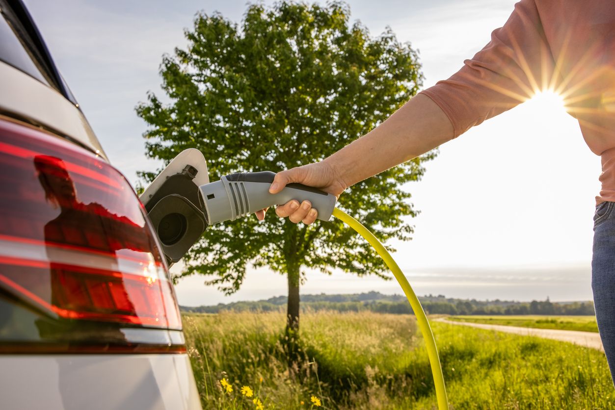Hand of woman inserting a power cord into an electric car for charging ecofriendly vehicle on green landscape
