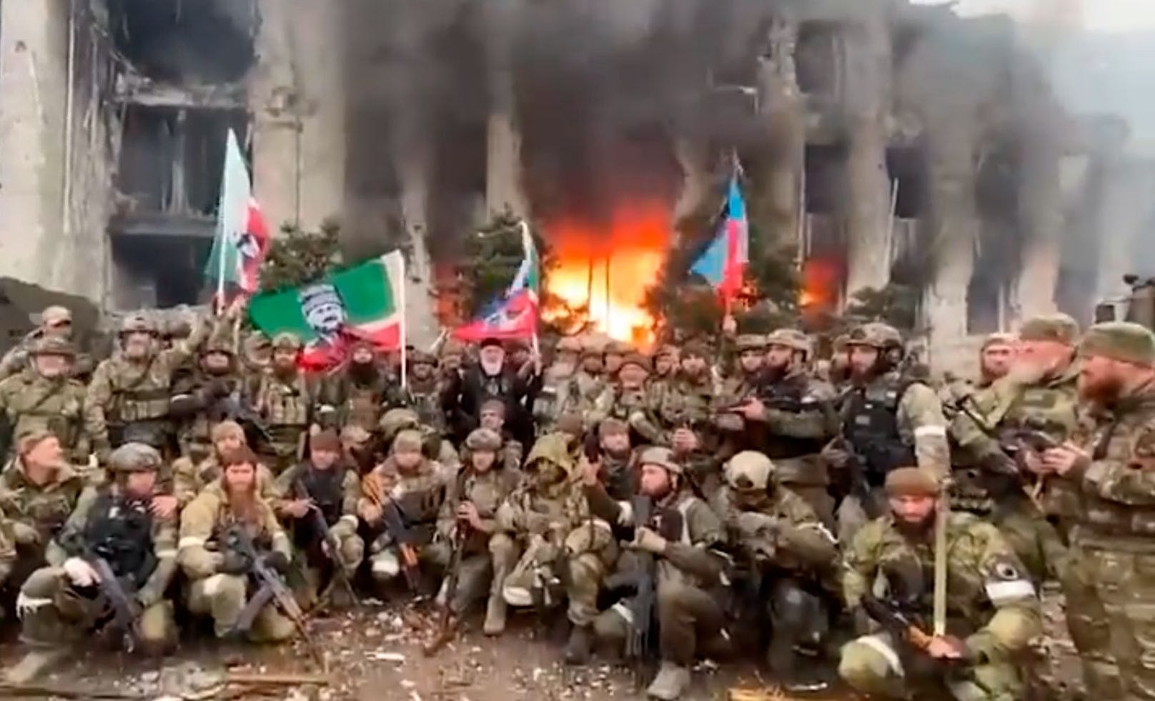 Tschetschenen feiern die Eroberung Mariupols vor einer brennenden Ruine.