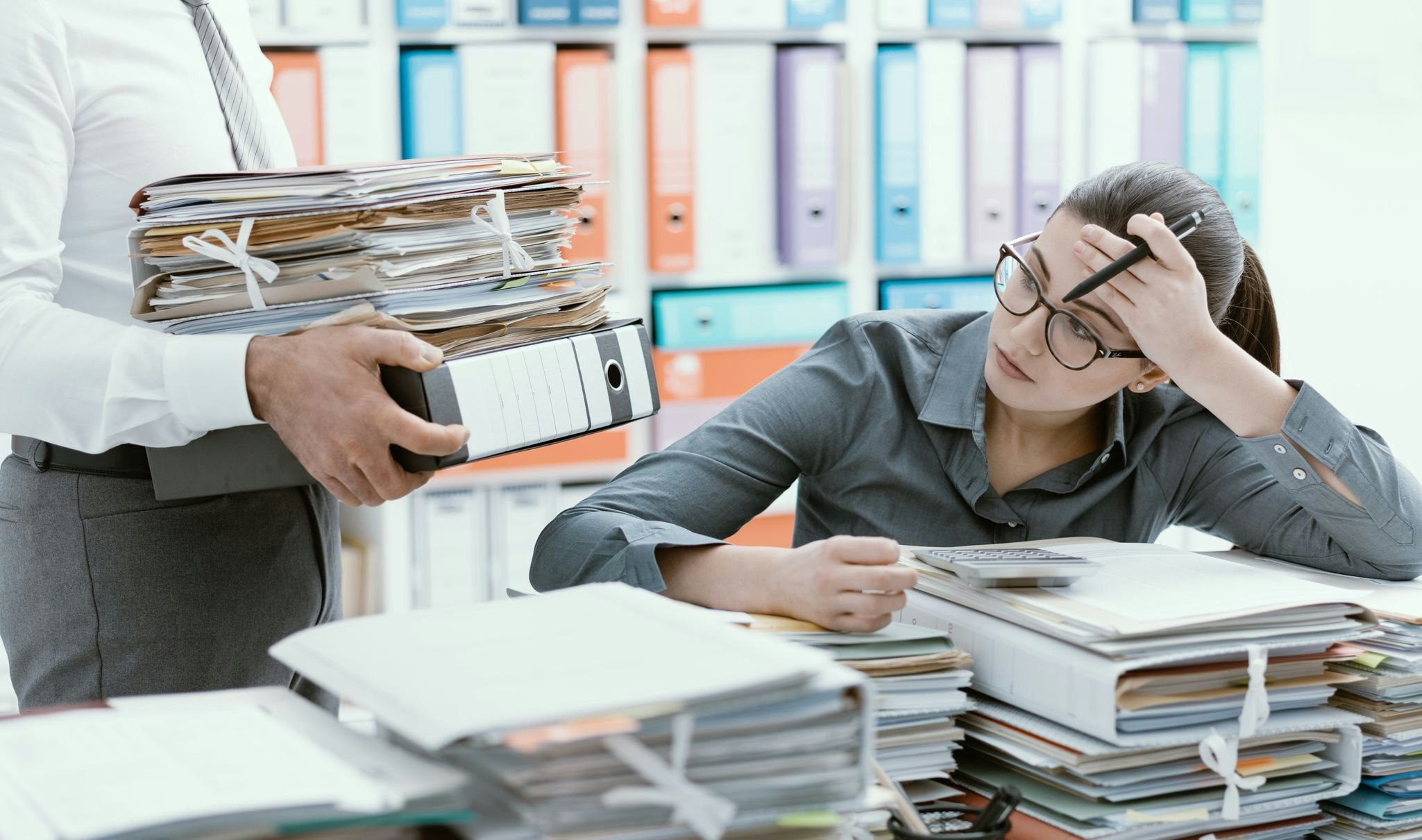 Young stressed secretary in the office overwhelmed by work and desk full of files, her boss is bringing more paperwork to her