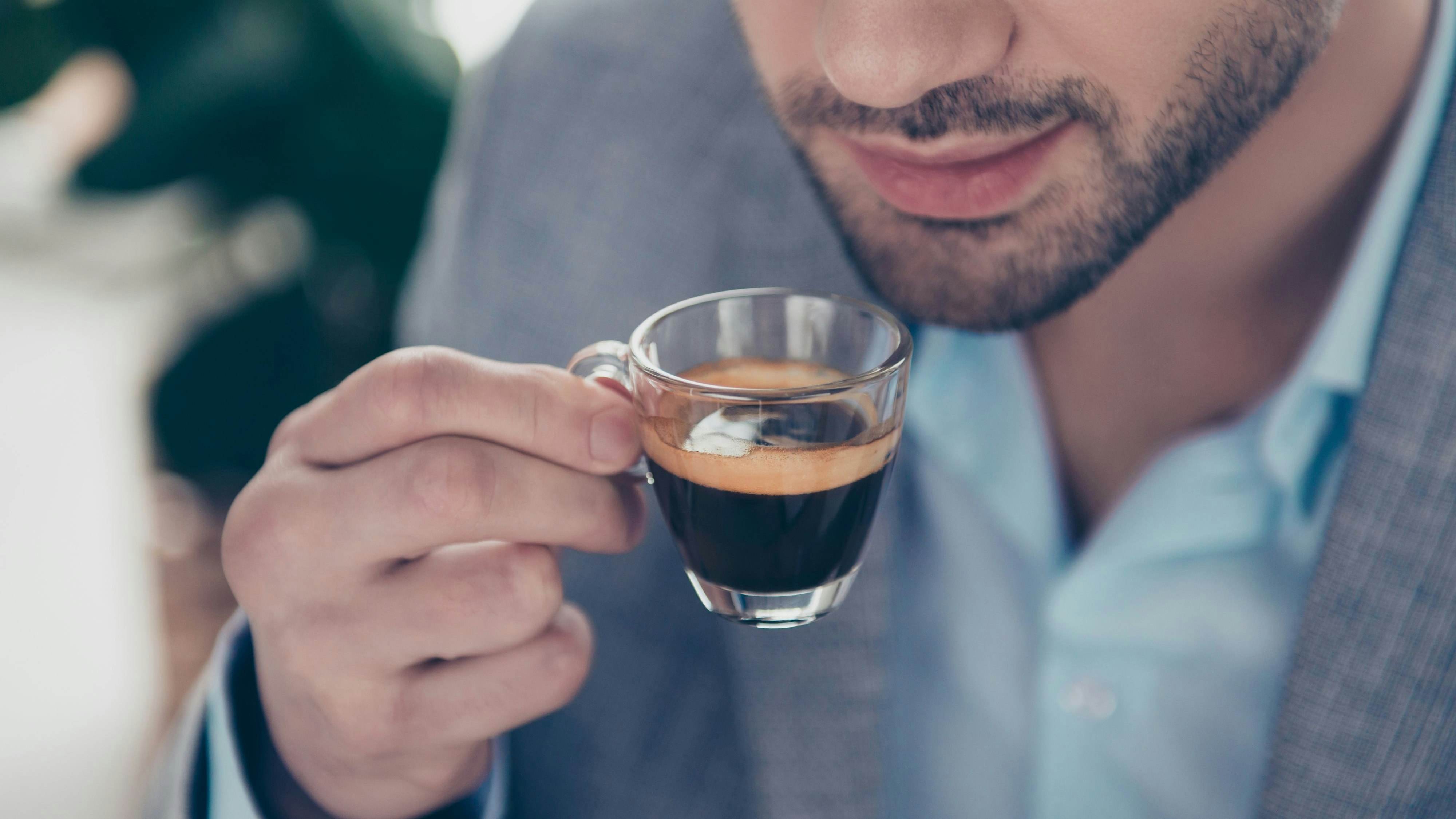 Cropped close up half face portrait of stylish attractive man holding small glass with espresso near mouth, every morning ritual before work in work place