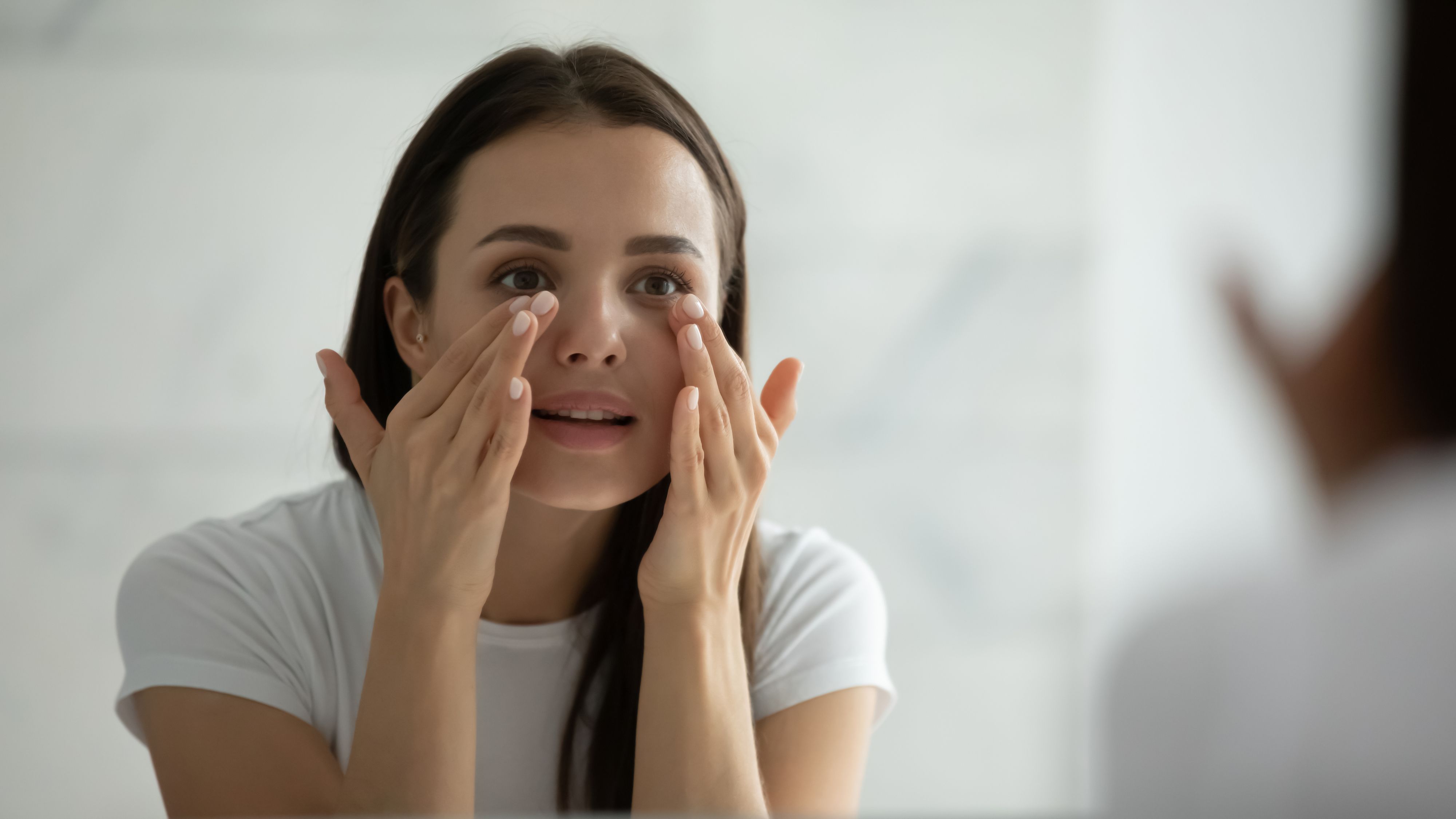 Close up smiling young woman wearing white t-shirt doing facial massage, applying moisturizing cream on under eye skin, looking in mirror, standing in bathroom, enjoying skincare procedure