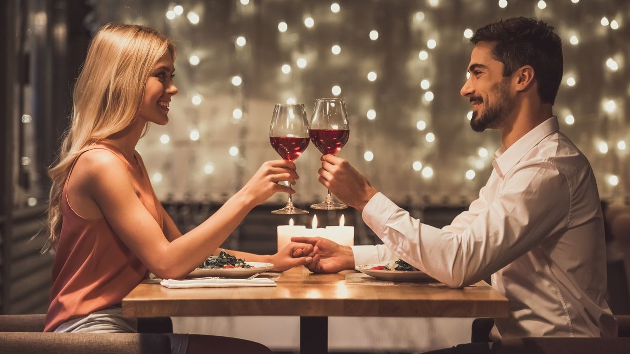 Beautiful young couple is looking at each other, clinking glasses of wine together and smiling during their date in a restaurant