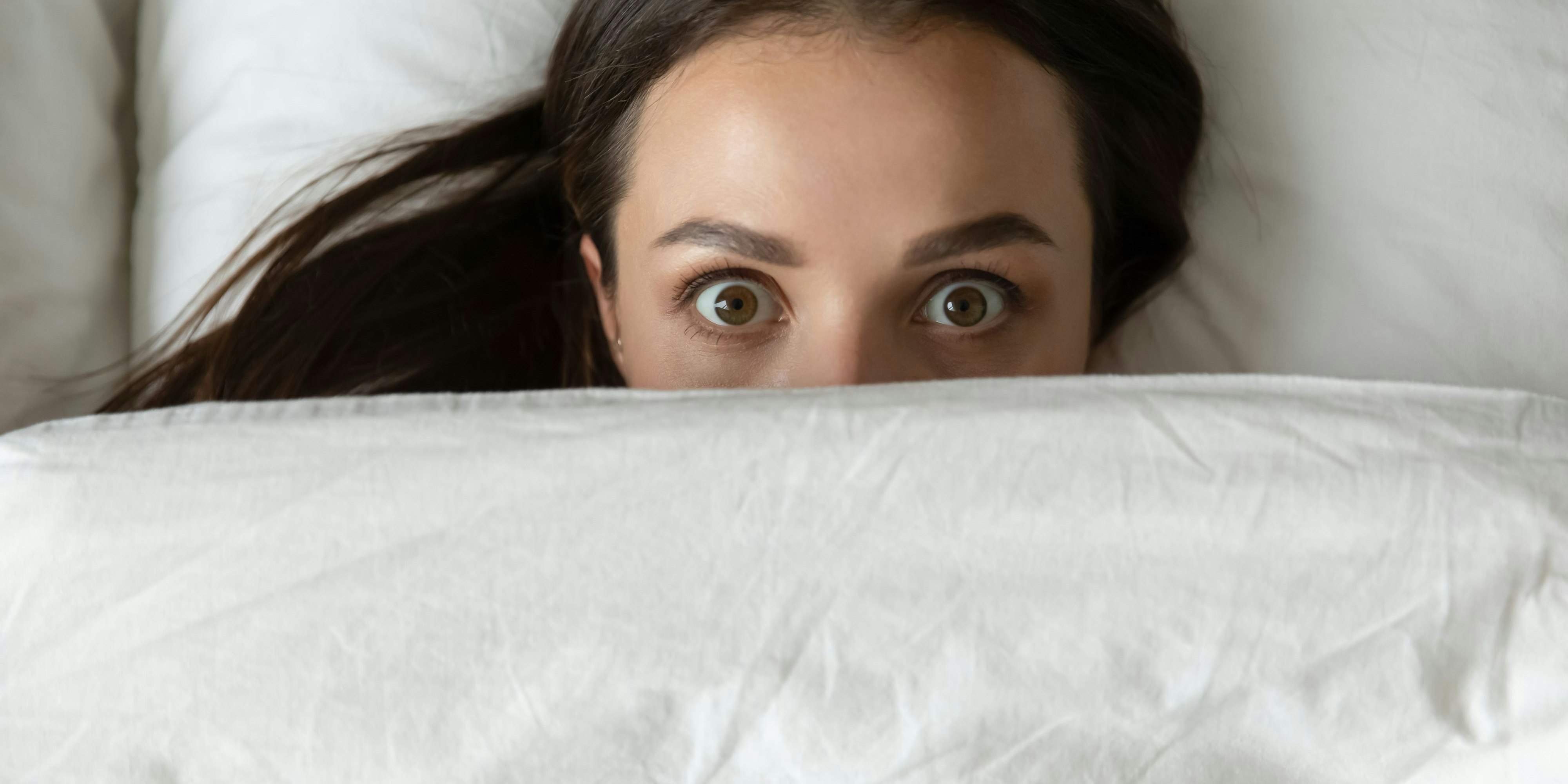 Close up top view funny young woman peeking from duvet, scared shocked female looking at camera, lying in bed with wide open eyes, afraid of nightmares, hiding under white warm blanket