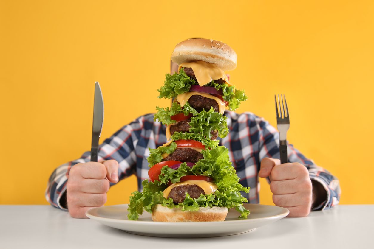 Hungry man with cutlery and huge burger at white table on yellow background