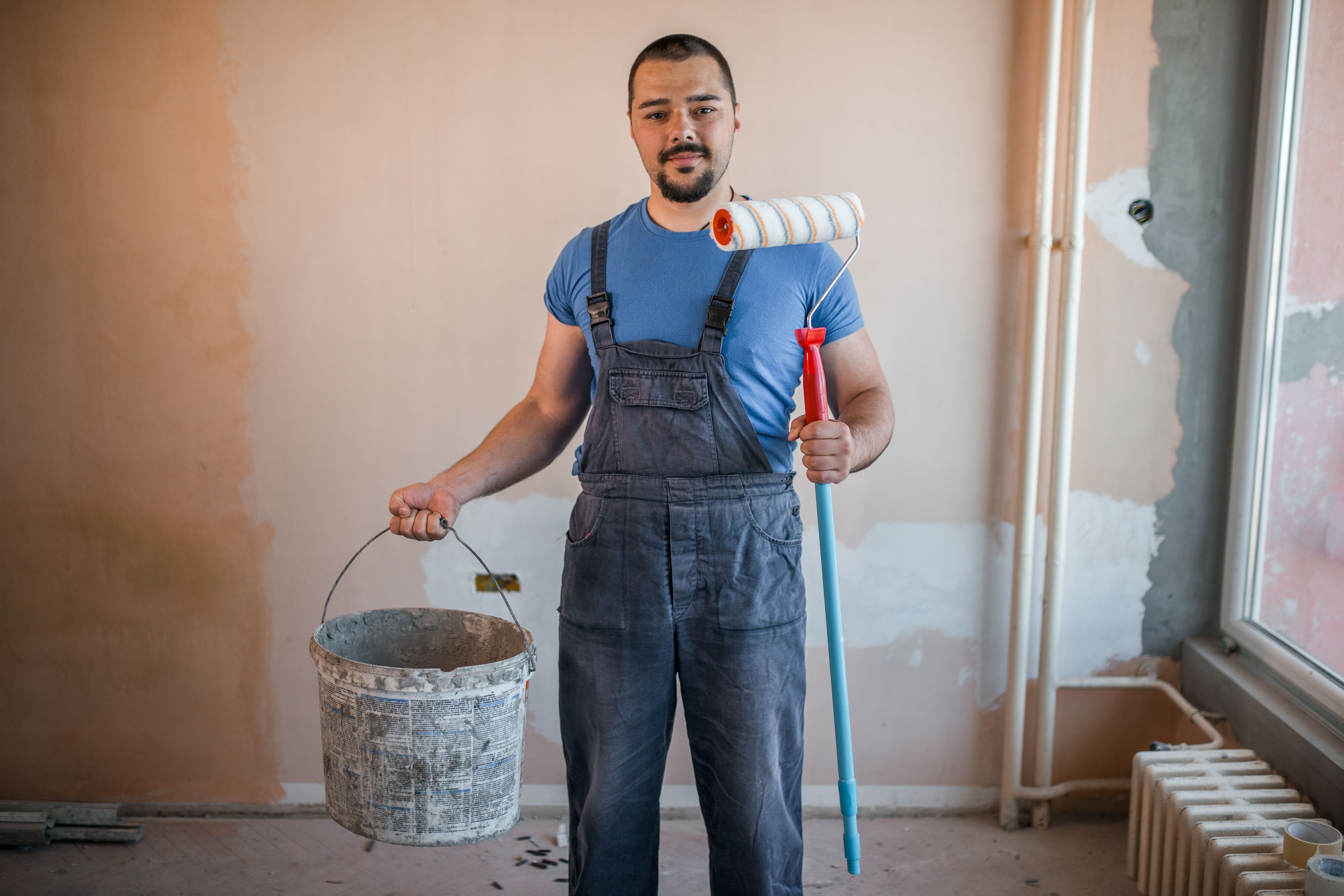 Handsome painter in coveralls holding paint roller and bucket