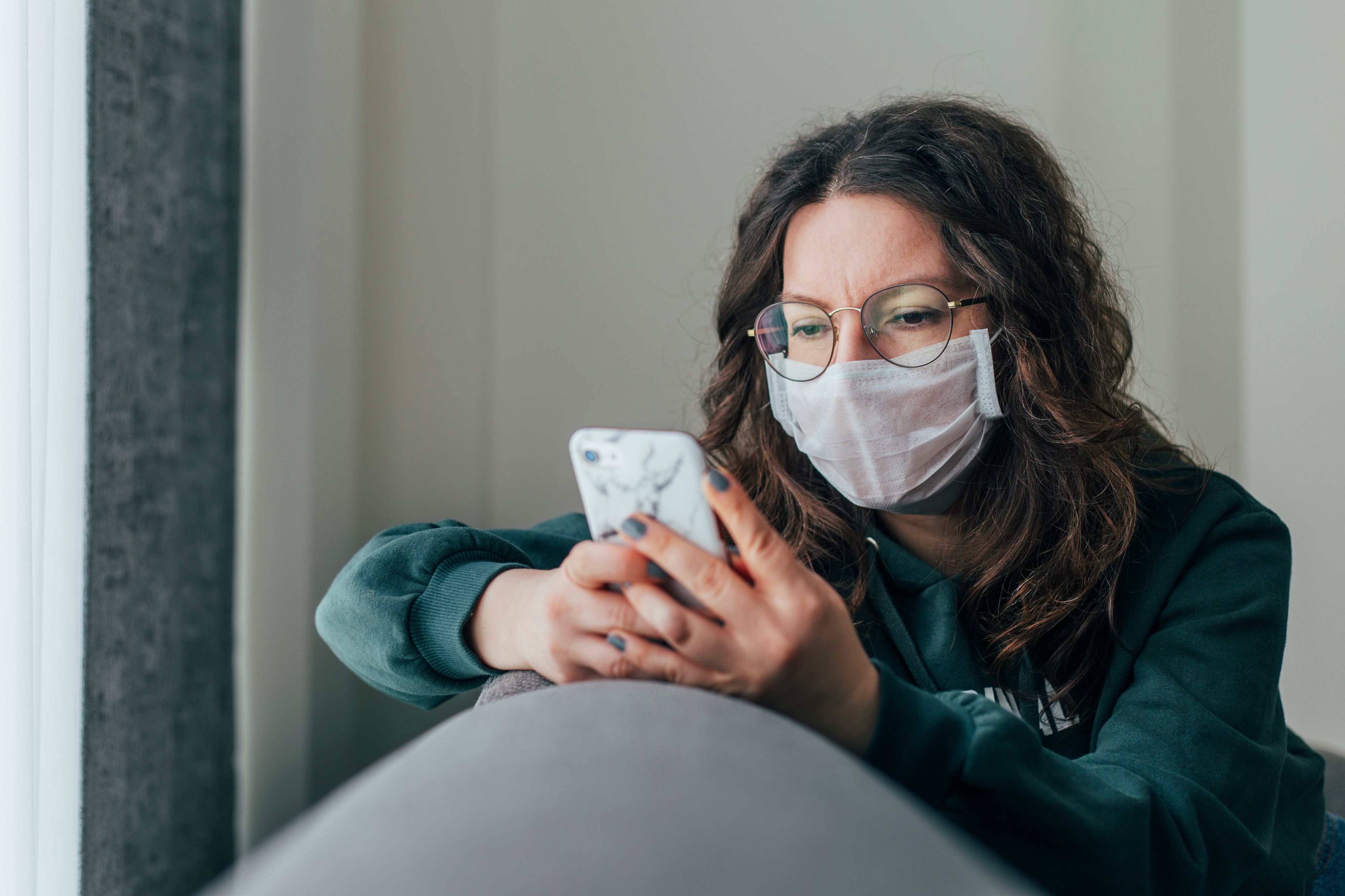 Young woman covering her face with surgical mask and using smartphone to find information about coronavirus spread on internet