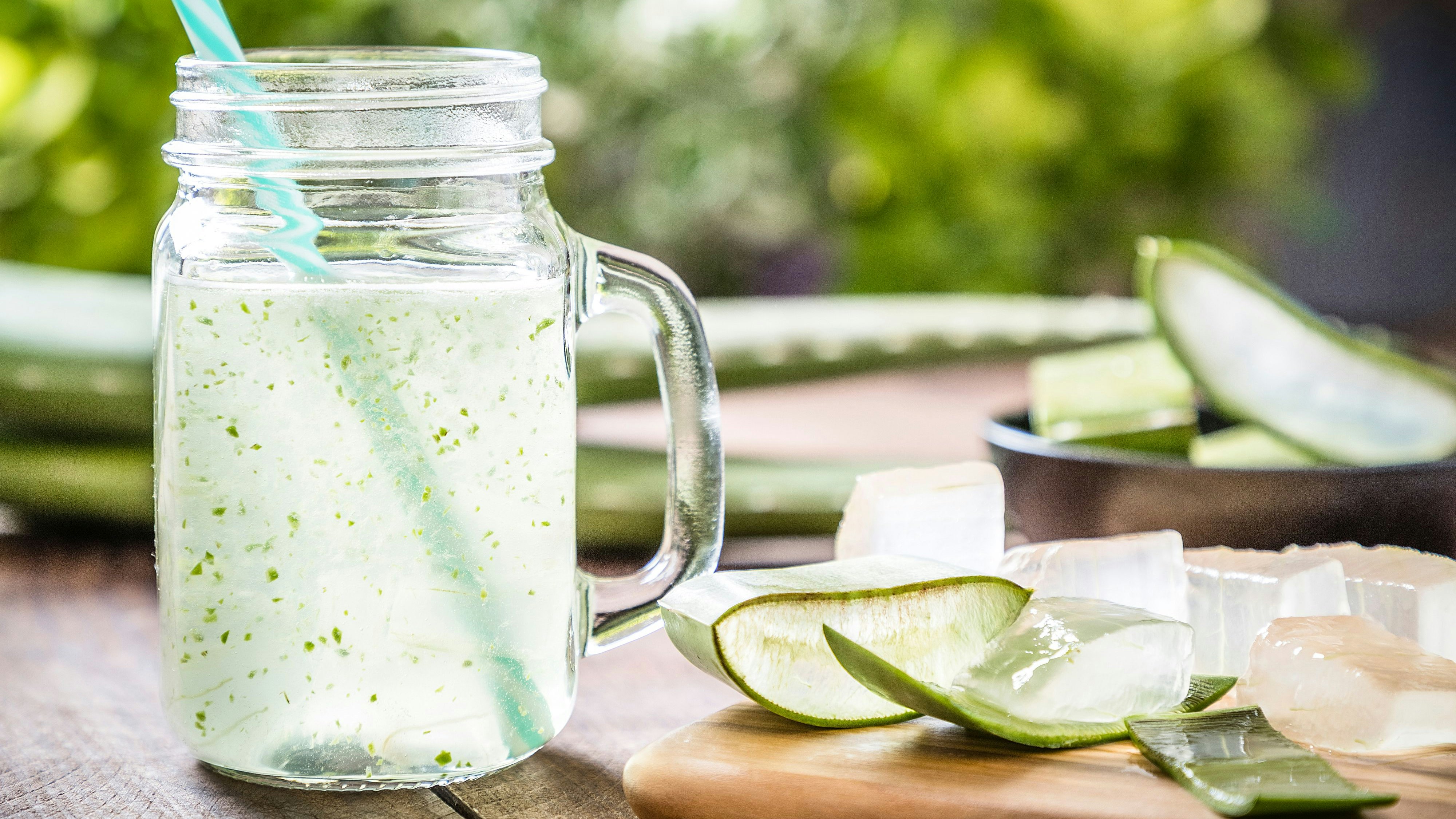 Front view of a drinking glass full of an aloe vera drink surrounded by some aloe vera crystals and sliced leaves. A black bowl full of sliced aloe vera leaves is defocused on the background. Predominant color are green and brown. Studio shot taken with Canon EOS 6D Mark II and Canon EF 24-105 mm f/4L