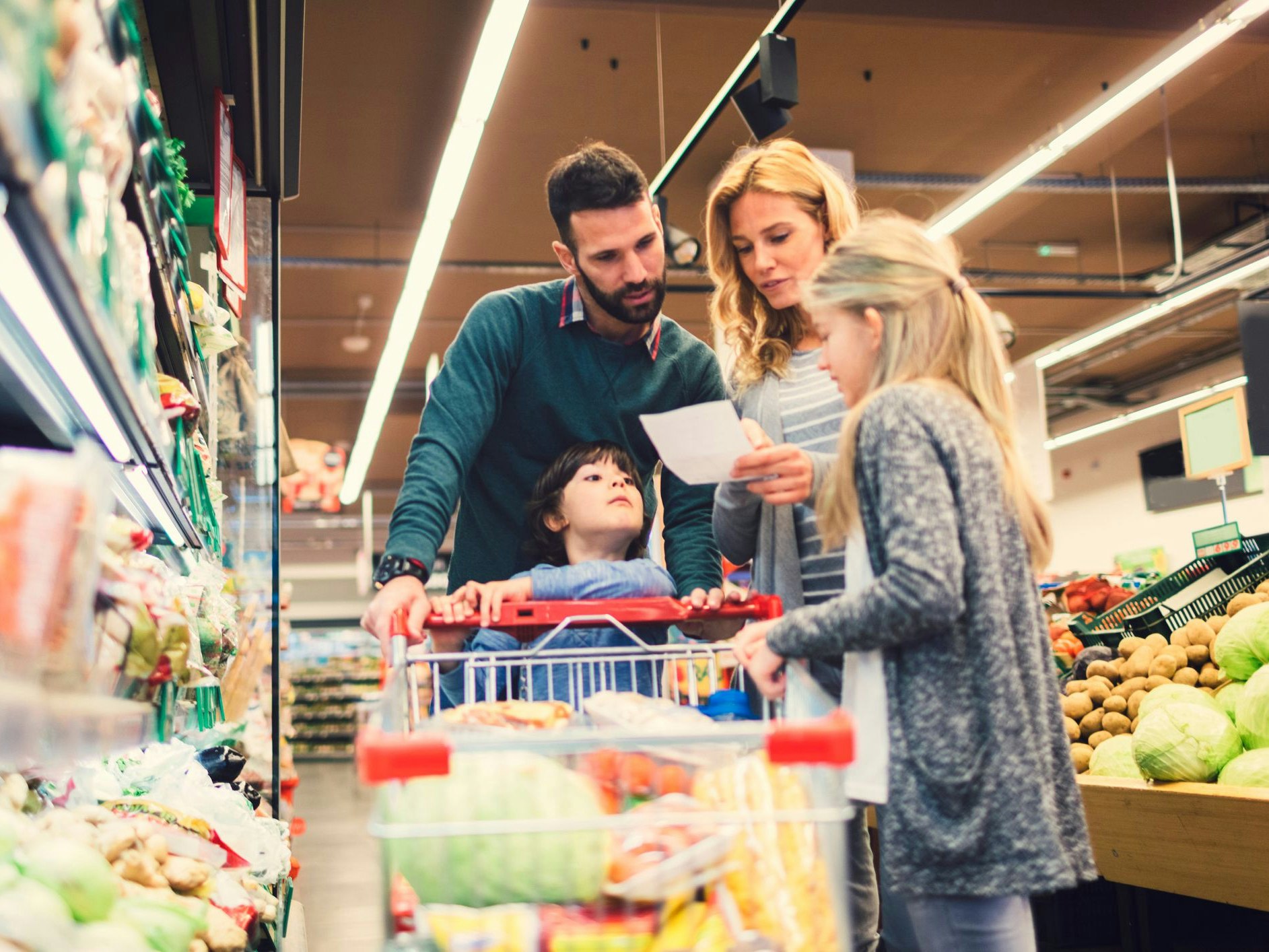 Cheerful family with two children shopping vegetables in supermarket. Holding shopping list and looking for groceries