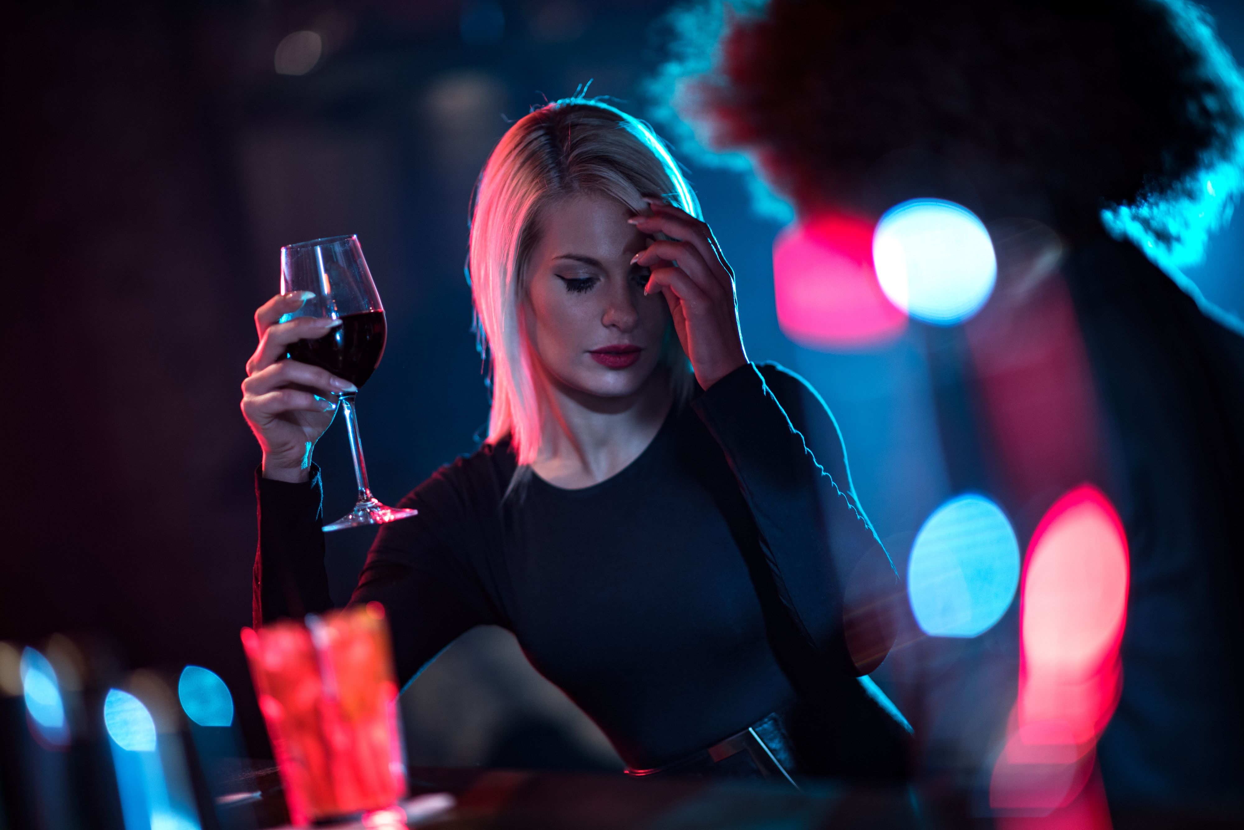 Blonde woman is holding a glass of red wine in the bar. Her female friend is caught up in lighting effect.