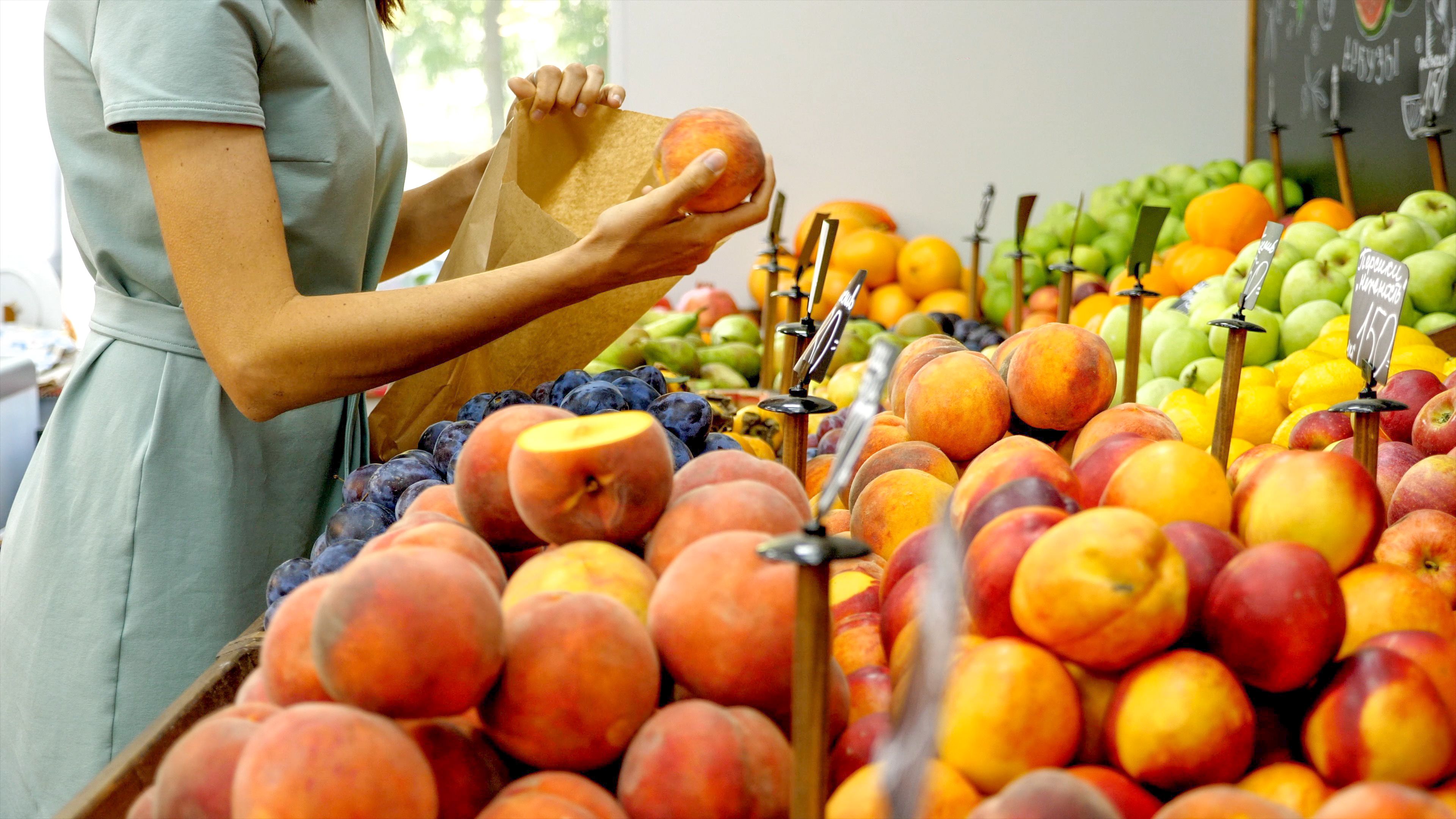 Young caucasian woman is choosing a fresh fruits at grocery store. Housewife in dress is taking ripe peaches from shelf and put it in paper bag.